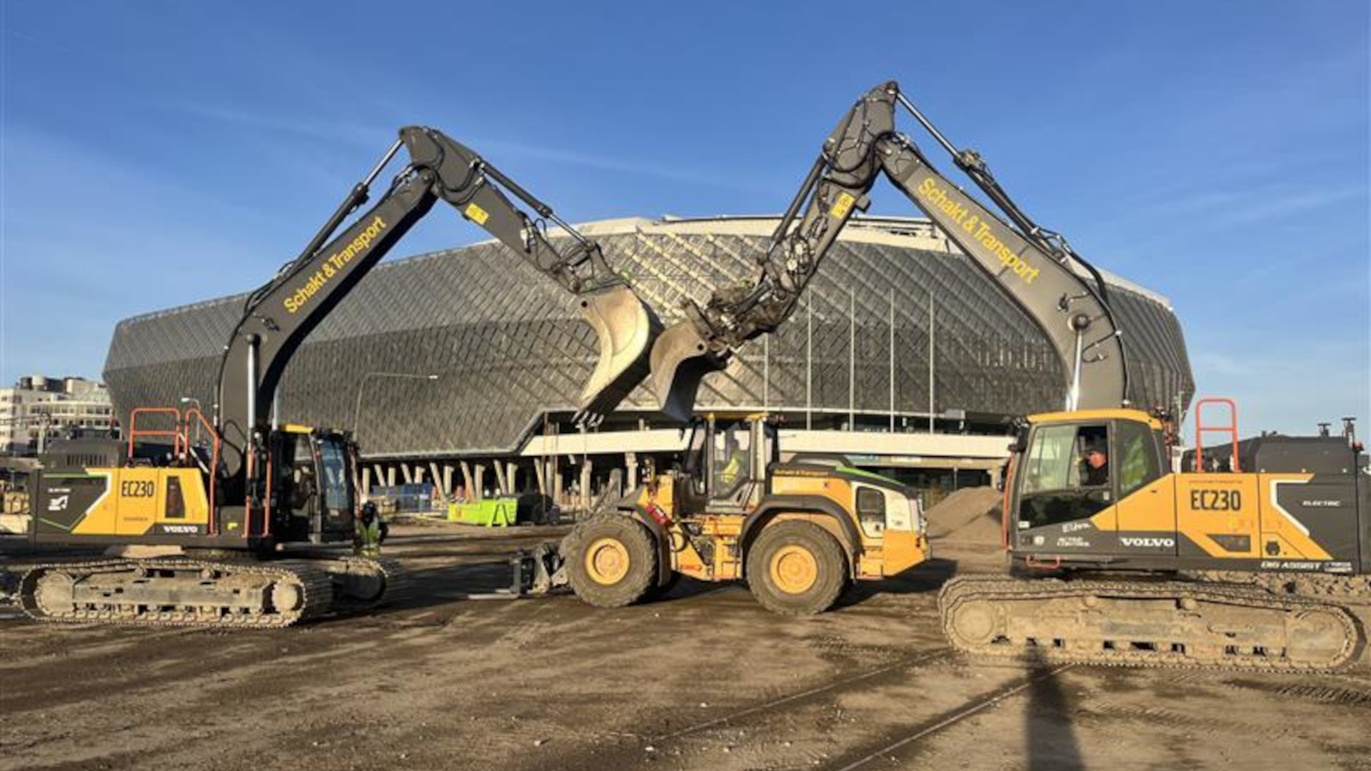 Eldrivna entreprenadmaskiner i samverkan på Slakthusområdet. Två grävmaskiner möter skoporna i luften på en byggarbetsplats, med en arena i bakgrunden.