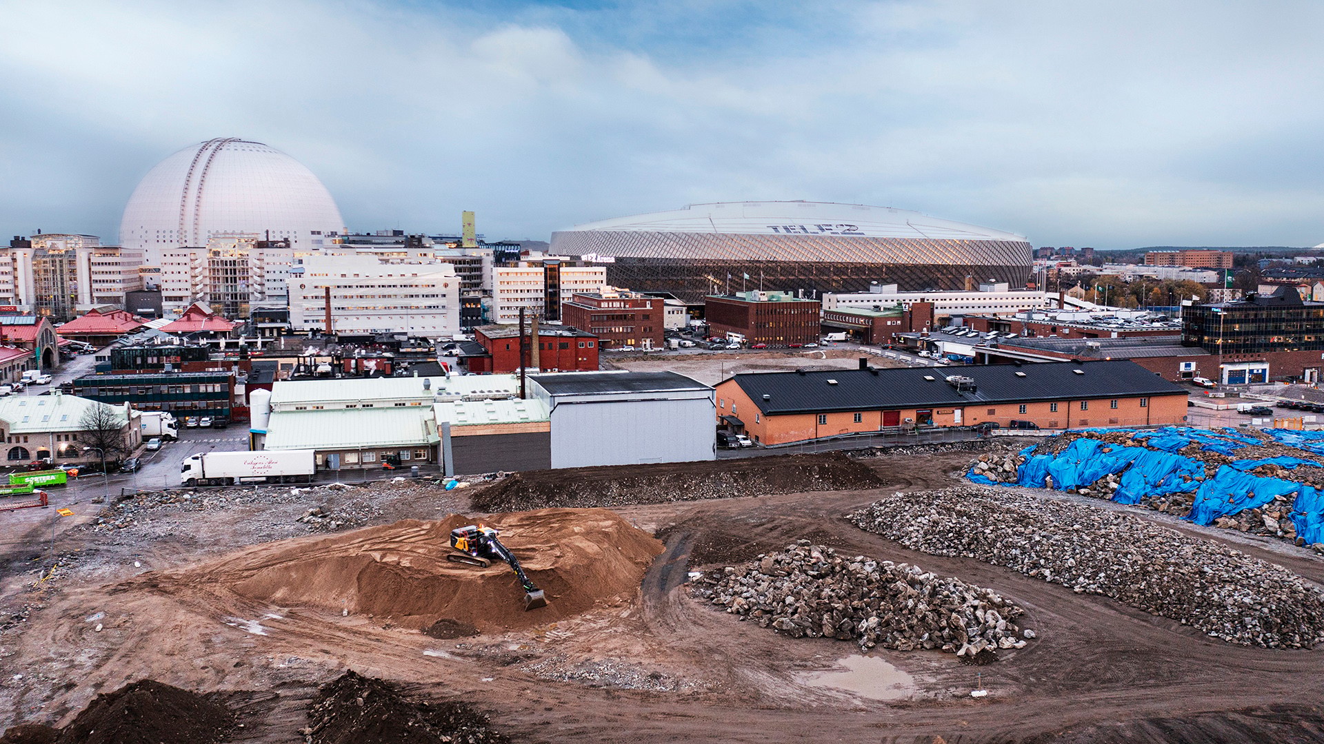 Stockholms stads höga kravställan om ett fossilbränsle- och emissionsfritt projekt har varit avgörande för detta banbrytande pilotprojekt. Här ses Volvos elektriska grävmaskin EC230 Electric. En byggarbetsplats med grävmaskin och stenhögar; i bakgrunden syns en stor arena och andra byggnader.