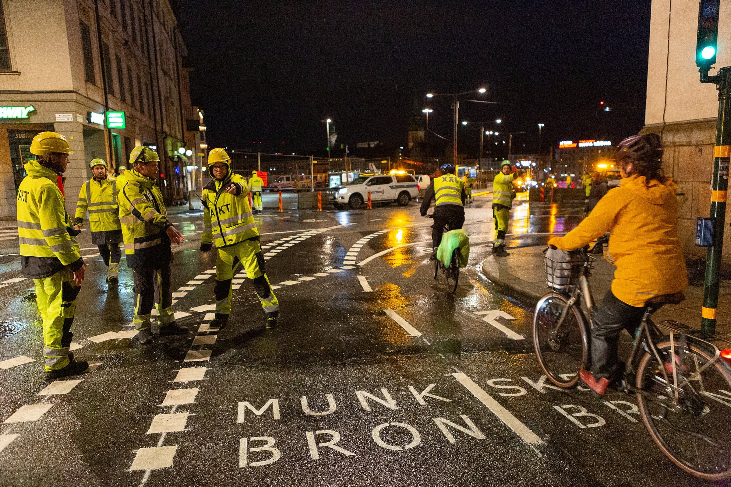 Arbetare i högsynlighetsutrustning hanterar nattligt vägarbete medan cyklister passerar på en våt gata.