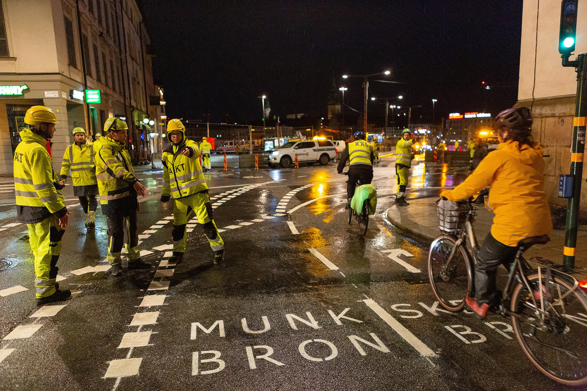 Anvisningar i gatan visar trafikanterna rätt. Arbetare i högsynlighetsutrustning hanterar nattligt vägarbete medan cyklister passerar på en våt gata.
