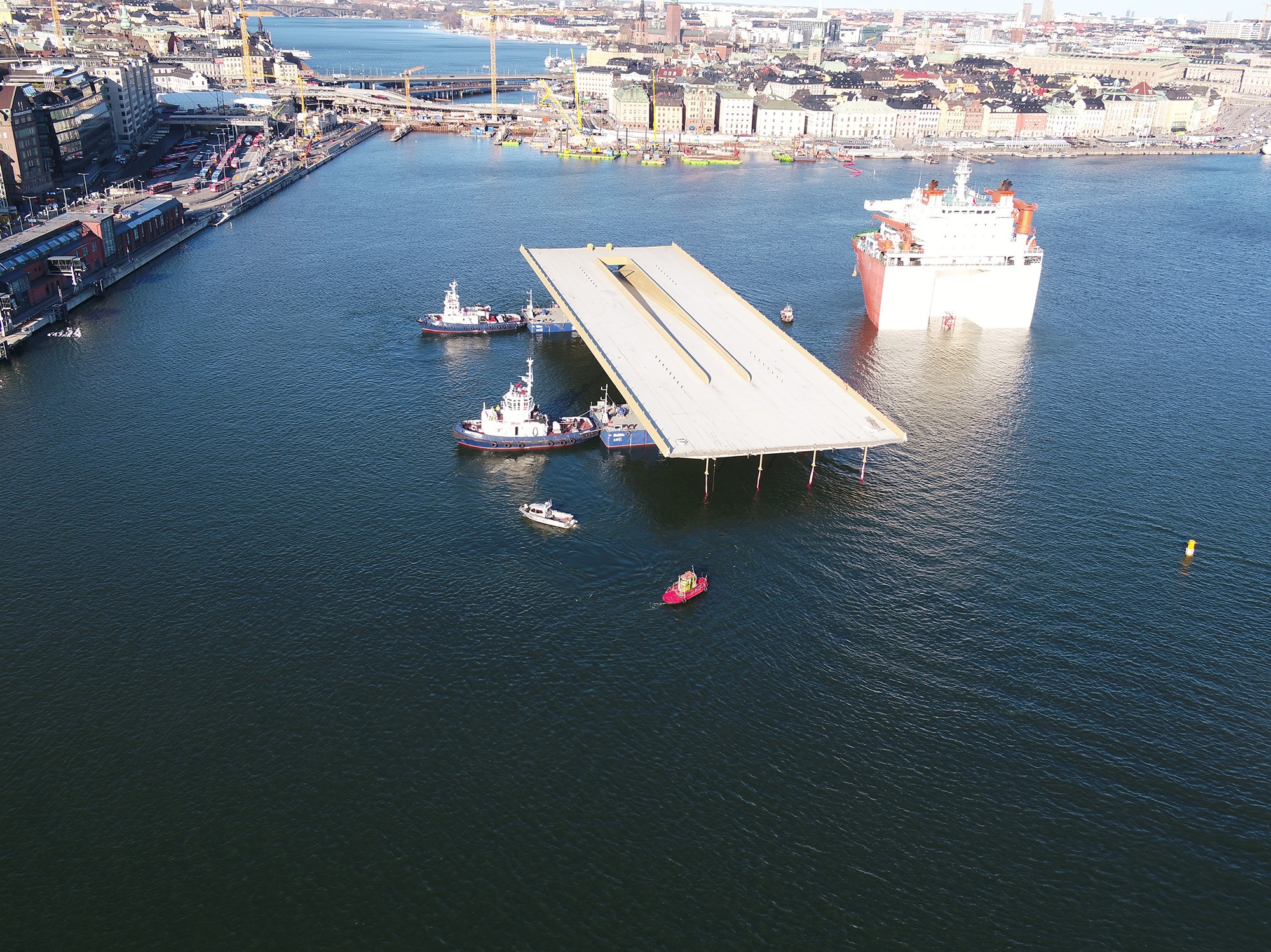Bogsering av bron in till kajen. Flygvy av en stor brosektion som transporteras av bogserbåtar i en hamn.