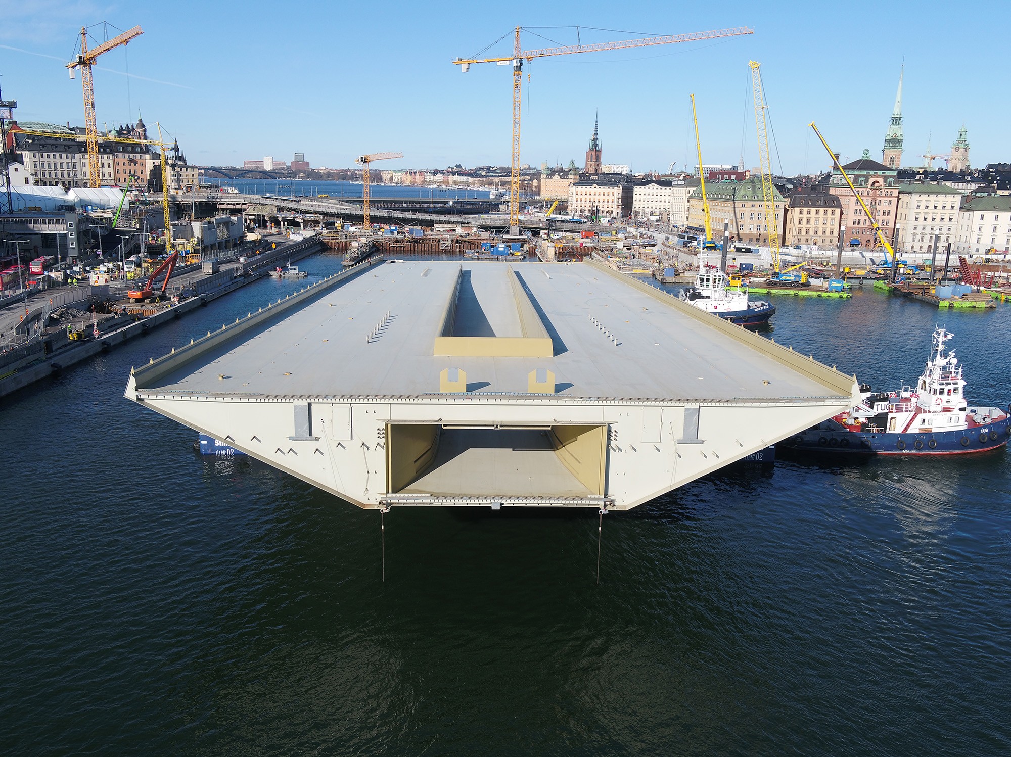 Bron är en rejäl pjäs med en beräknad livslängd på minst 120 år. En stor brosektion transporteras av bogserbåtar i en stadsbild med kranar och byggnader.