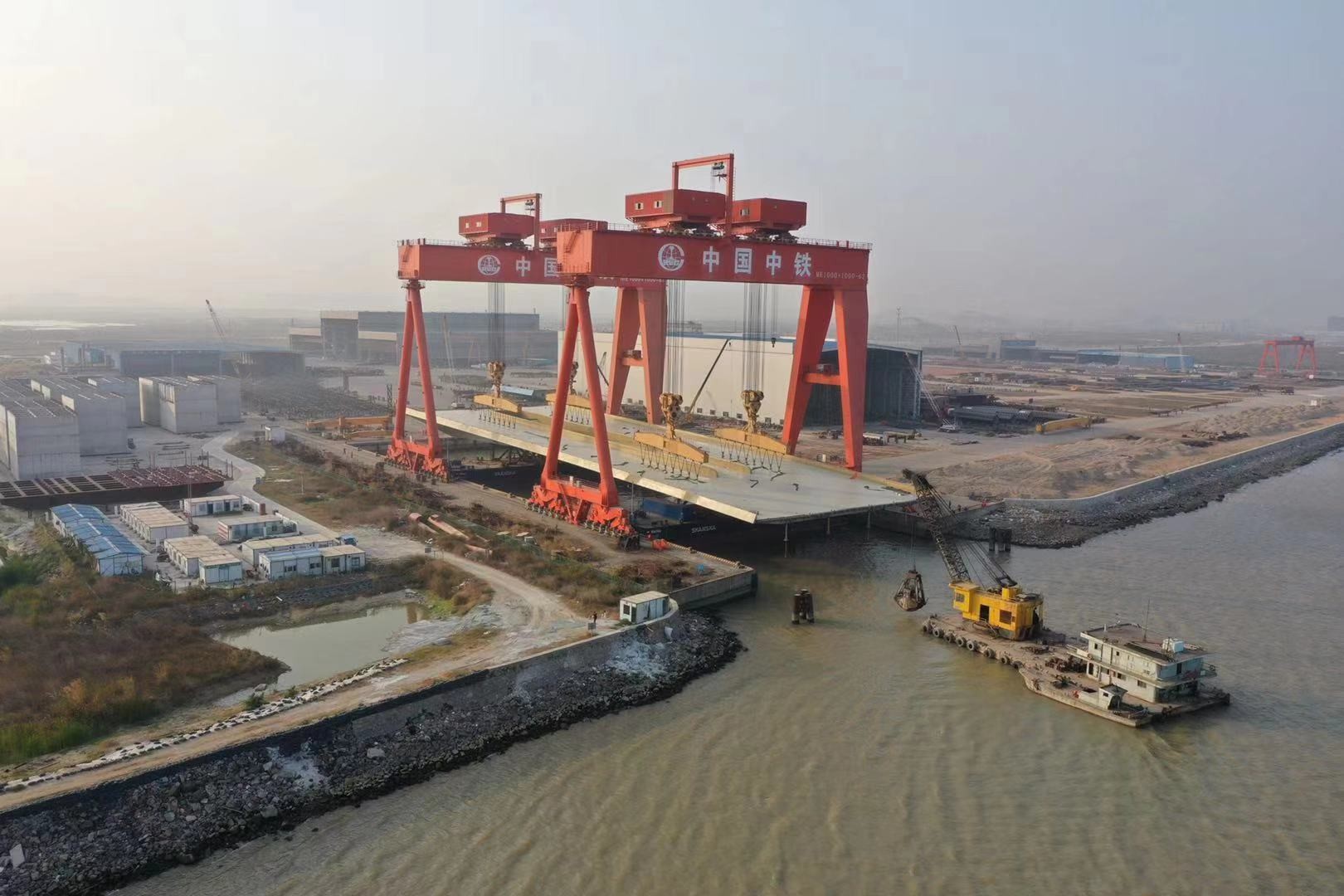 Den färdiga bron lyftes över till två pontoner med hjälp av två traverskranar som tar 2000 ton vardera. Källa för bilder: Foster+Partners Perry Ip Stora röda portalkranar över en betongstruktur nära en byggarbetsplats vid vattnet.