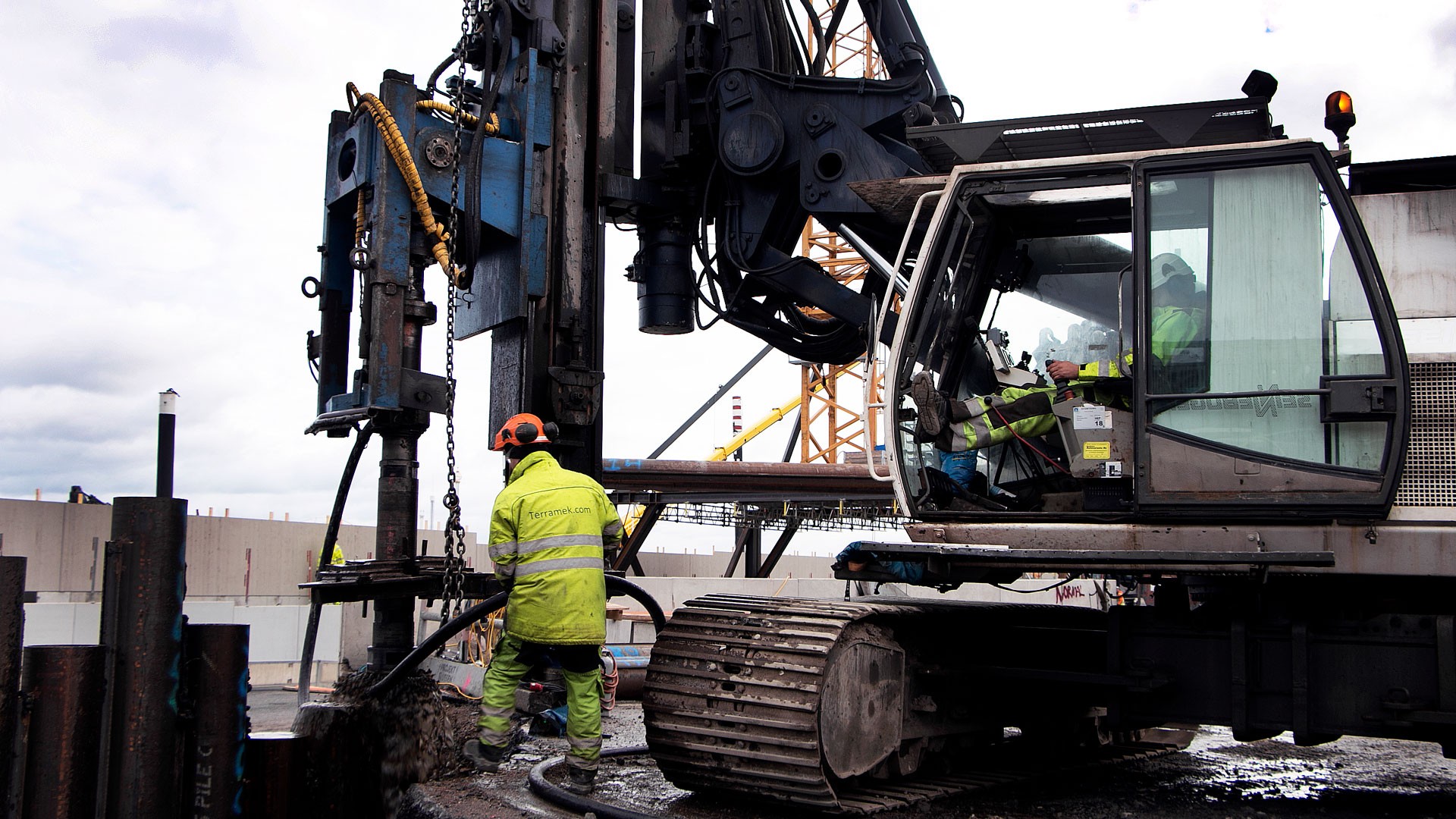 Det sker mycket pålning under projektets gång. Byggnadsarbetare som hanterar en stor pålningsrigg på en lerig byggarbetsplats.