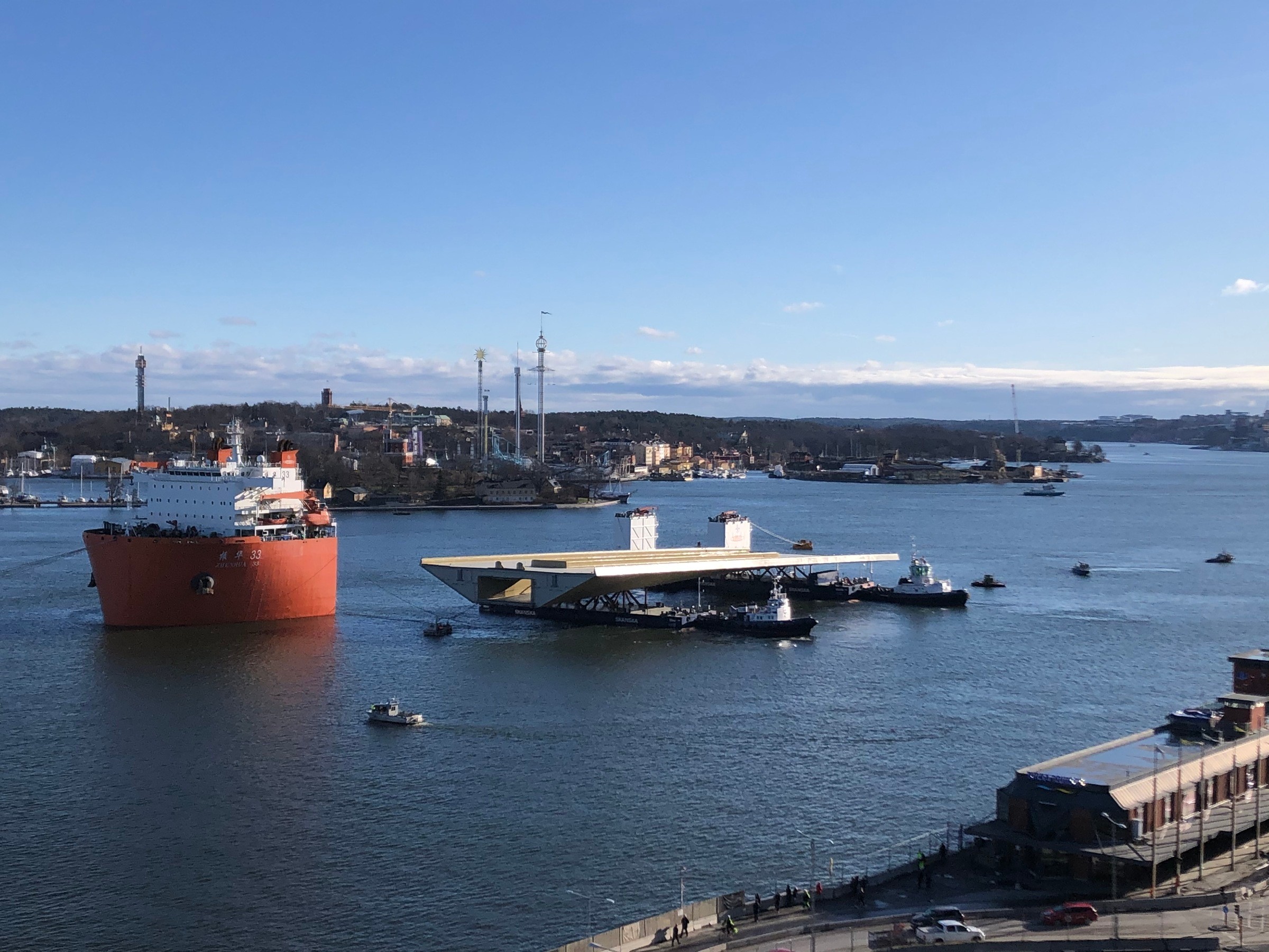 Fartyget är sänkt till lägsta nivå och förbereds för att bogsera bron och pontonerna av fartyget och till kaj Ett stort skepp navigerar i en hamn, eskorterat av bogserbåtar, under en klarblå himmel.
