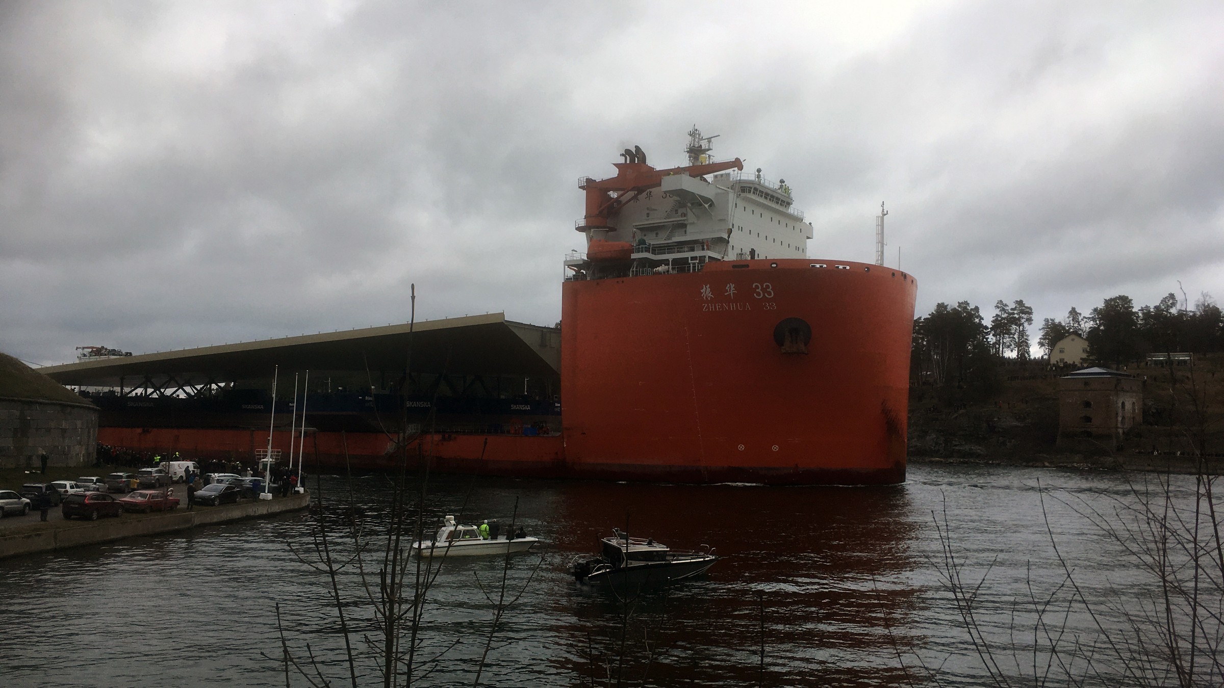 Fartyget med nya Slussenbron passerar Oxdjupet, Rindö, på väg in mot Stockholm 11 mars 2020. Stort rött lastfartyg "Zhen Hua 33" lagt till nära en marina under en molnig himmel.