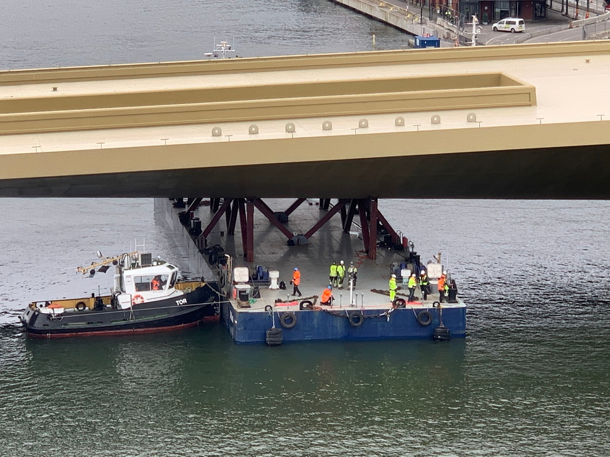 Förflyttningen av bron genomförs med största noggrannhet. En pråm som transporterar byggnadsarbetare och utrustning är placerad under en stor bro över vatten.