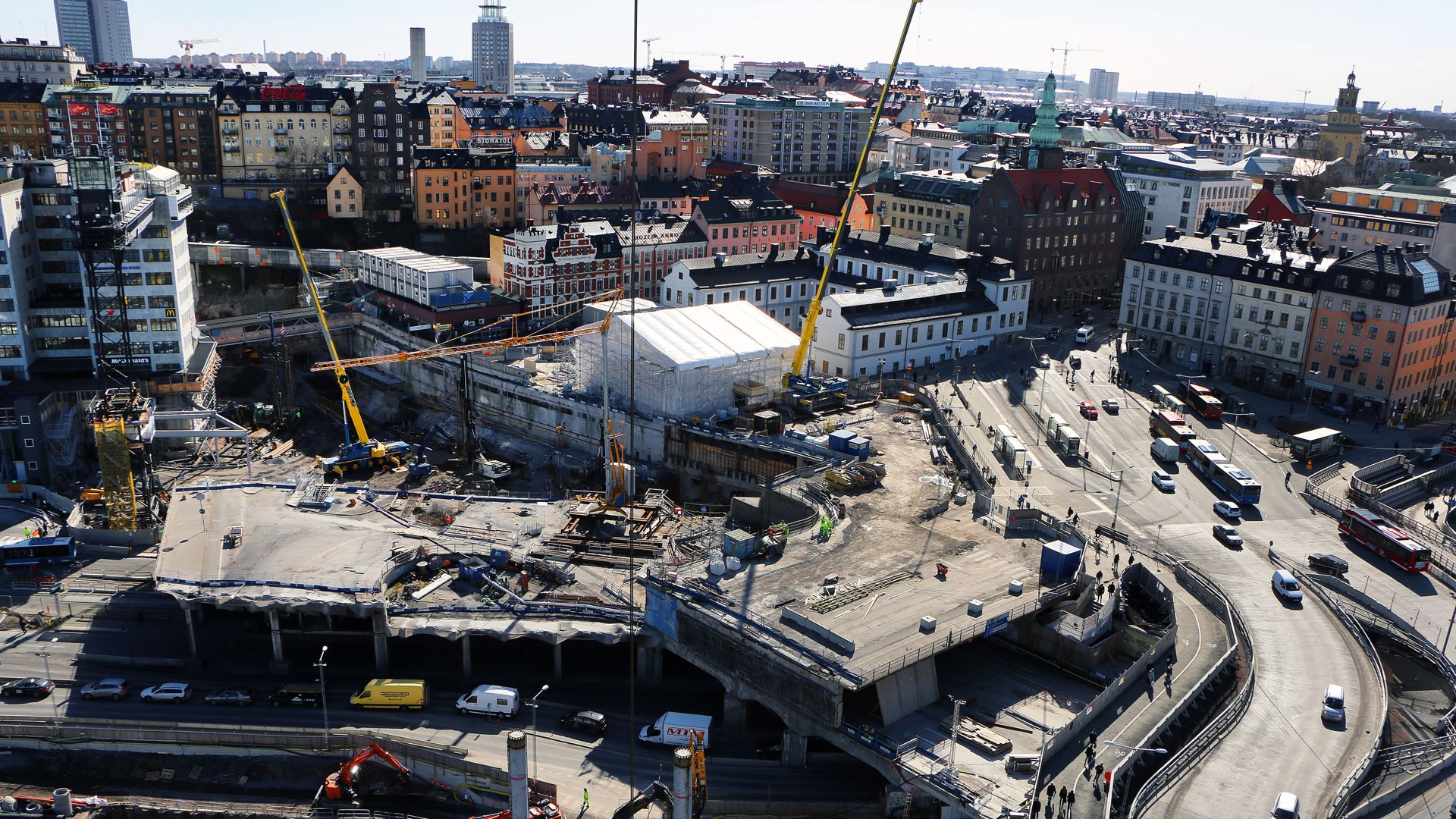 Från tornkranen på Slussenprojektet breder en fantastisk utsikt ut sig. Flygvy över stadsbyggplats med kranar och fordon bland historiska stadsbyggnader.