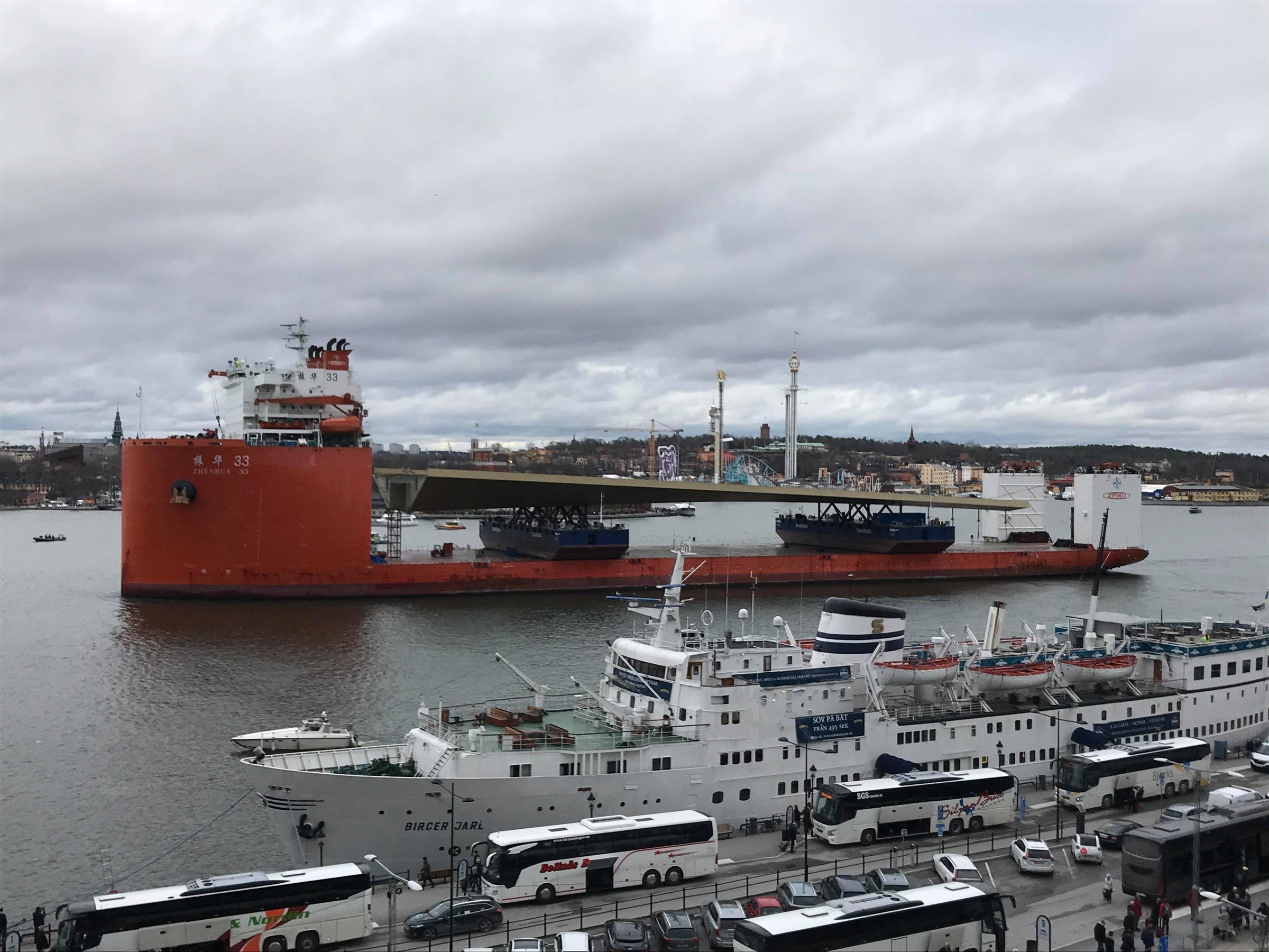 Nu är den här. Välkommen Stockholms gyllene Slussenbro! Stort rött lastfartyg som transporterar industriutrustning passerar förtöjda vita passagerarfartyg i molnig hamn.