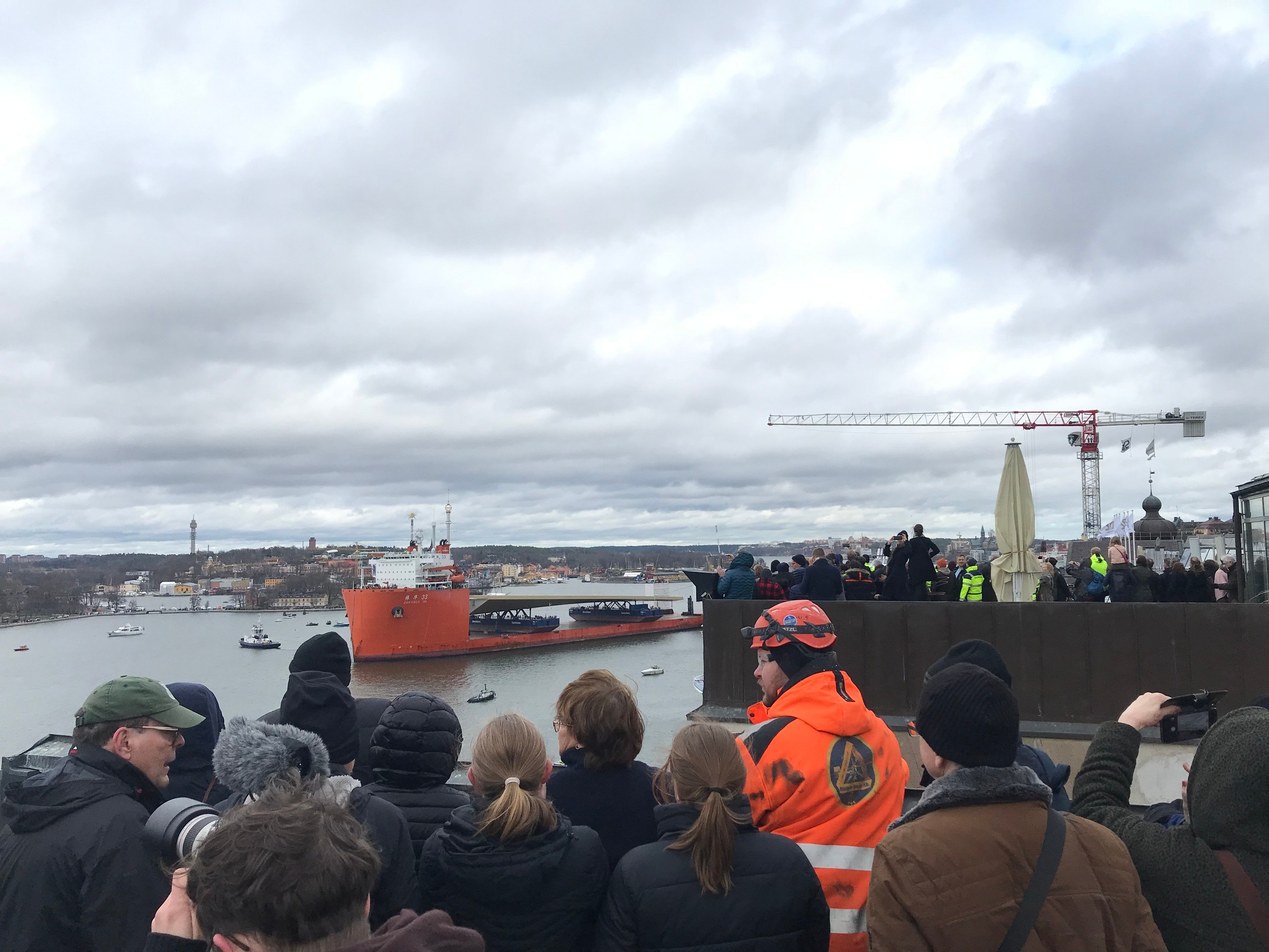 Nya slussenbron är nära sitt mål, här i inloppet till Stockholm. En folkmassa tittar på ett stort orange skepp i en hamn, med kranar och byggnader i bakgrunden.
