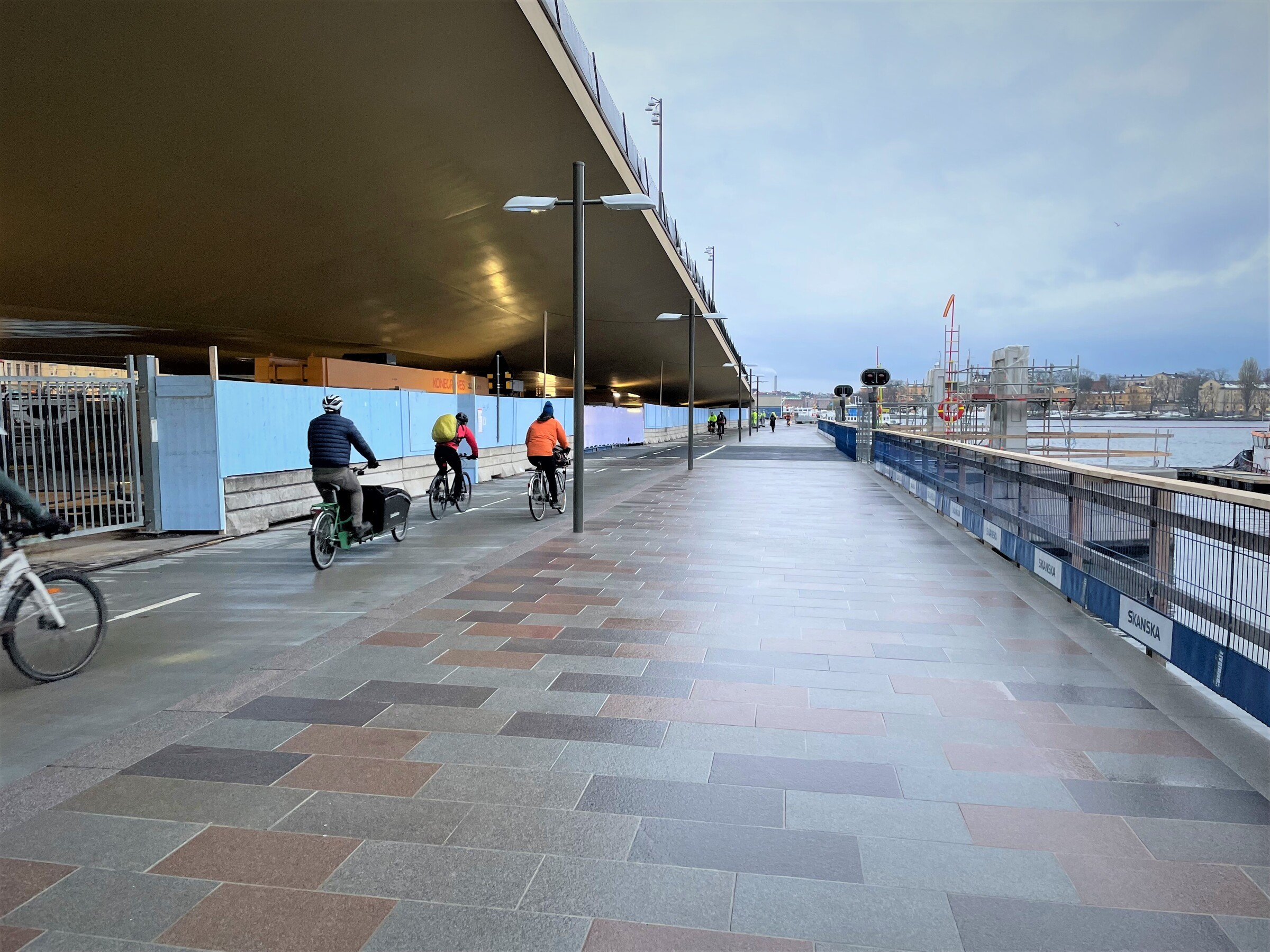 Östra lågbron öppnade för gång- och cykeltrafik i slutet av november 2022. Cyklister på cykelbanan som går under den guldgula Slussenbron, till höger en plattbelagd gångväg utmed Saltsjön. I bakgrunden skymtar siluetten av Gamla stan.