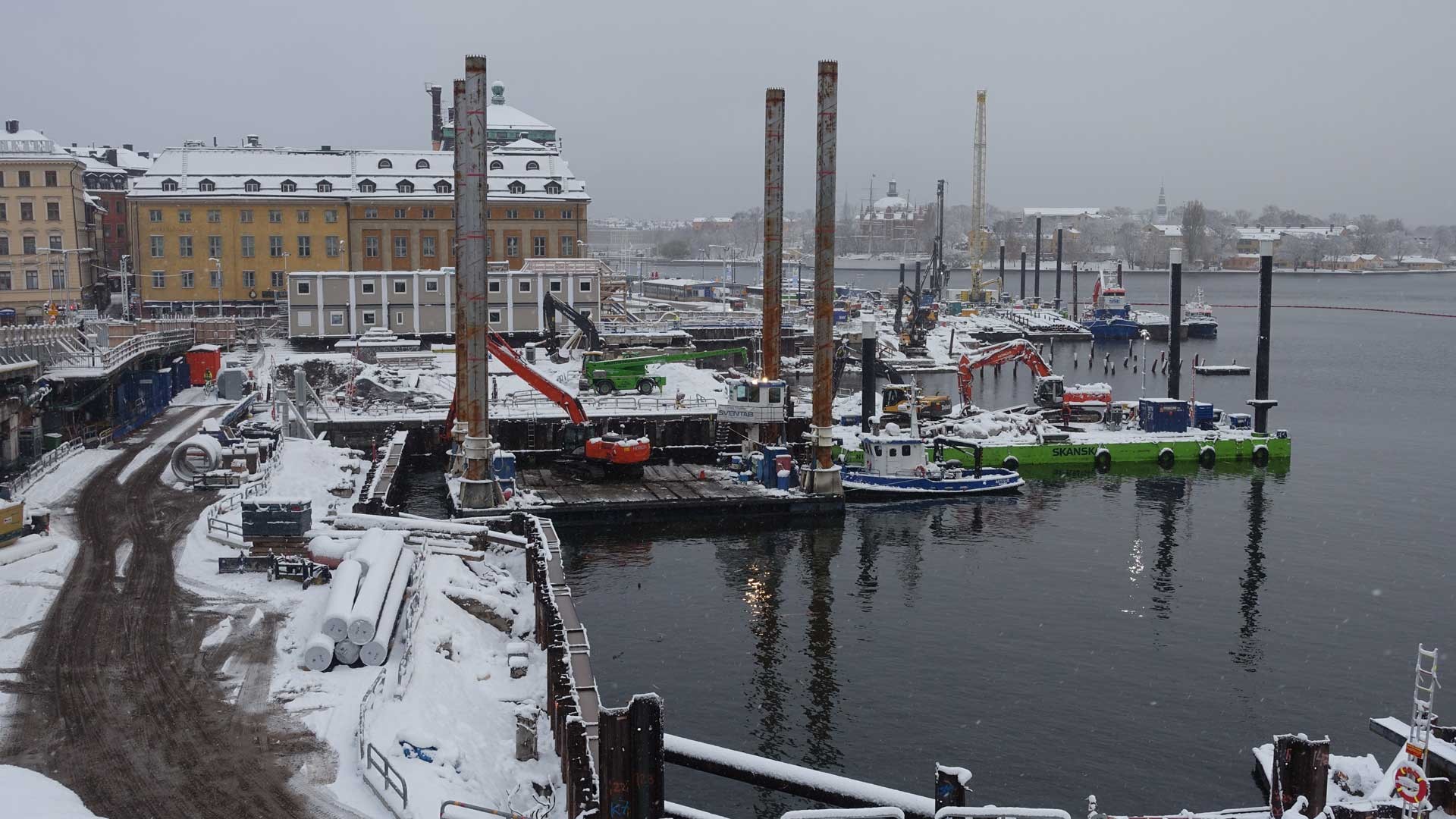 Pontoner under vintern Snötäckt byggarbetsplats vid en flod, med kranar och båtar nära historiska byggnader.