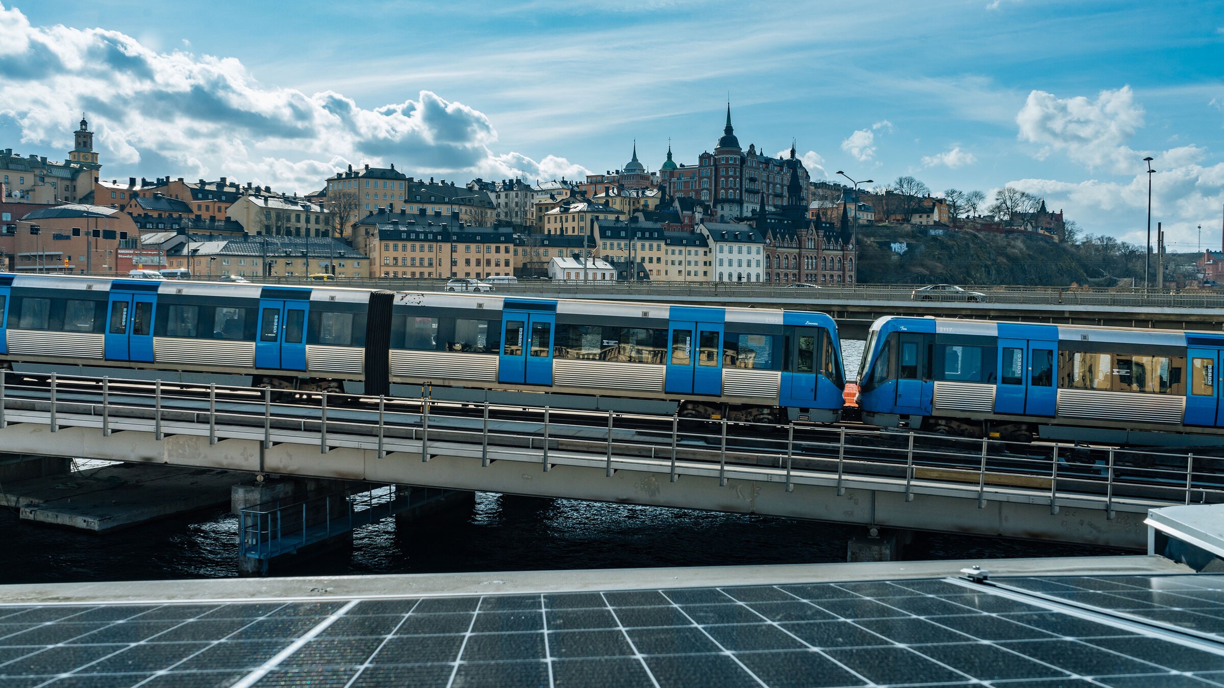 Projektet med solceller på byggbodarnas tak stöds av Bra Miljöval och kan på sikt minska både effektuttag och klimatpåverkan. Tåg som korsar en bro med en naturskön utsikt över Stockholms historiska silhuett under en molnig himmel.