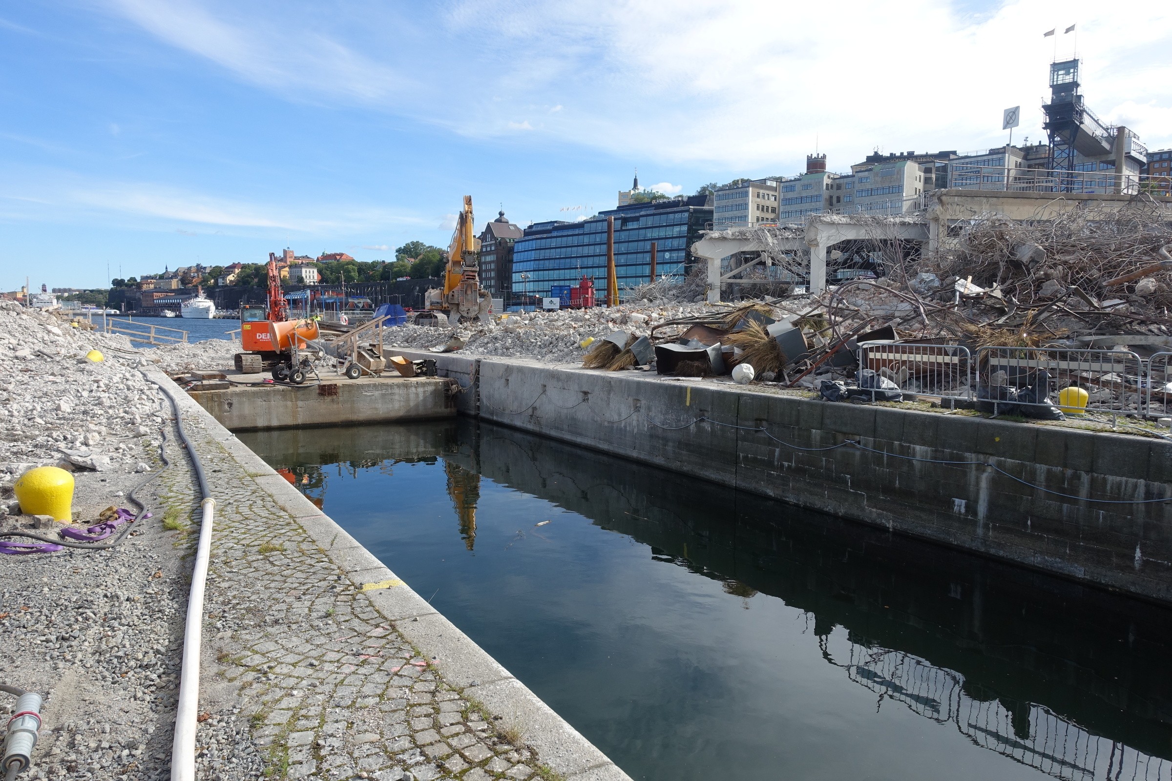 Rivning vid Slusskanalen, september 2016 Byggarbetsplats med bråte och maskiner nära en strandlinje, omgiven av stadens byggnader.