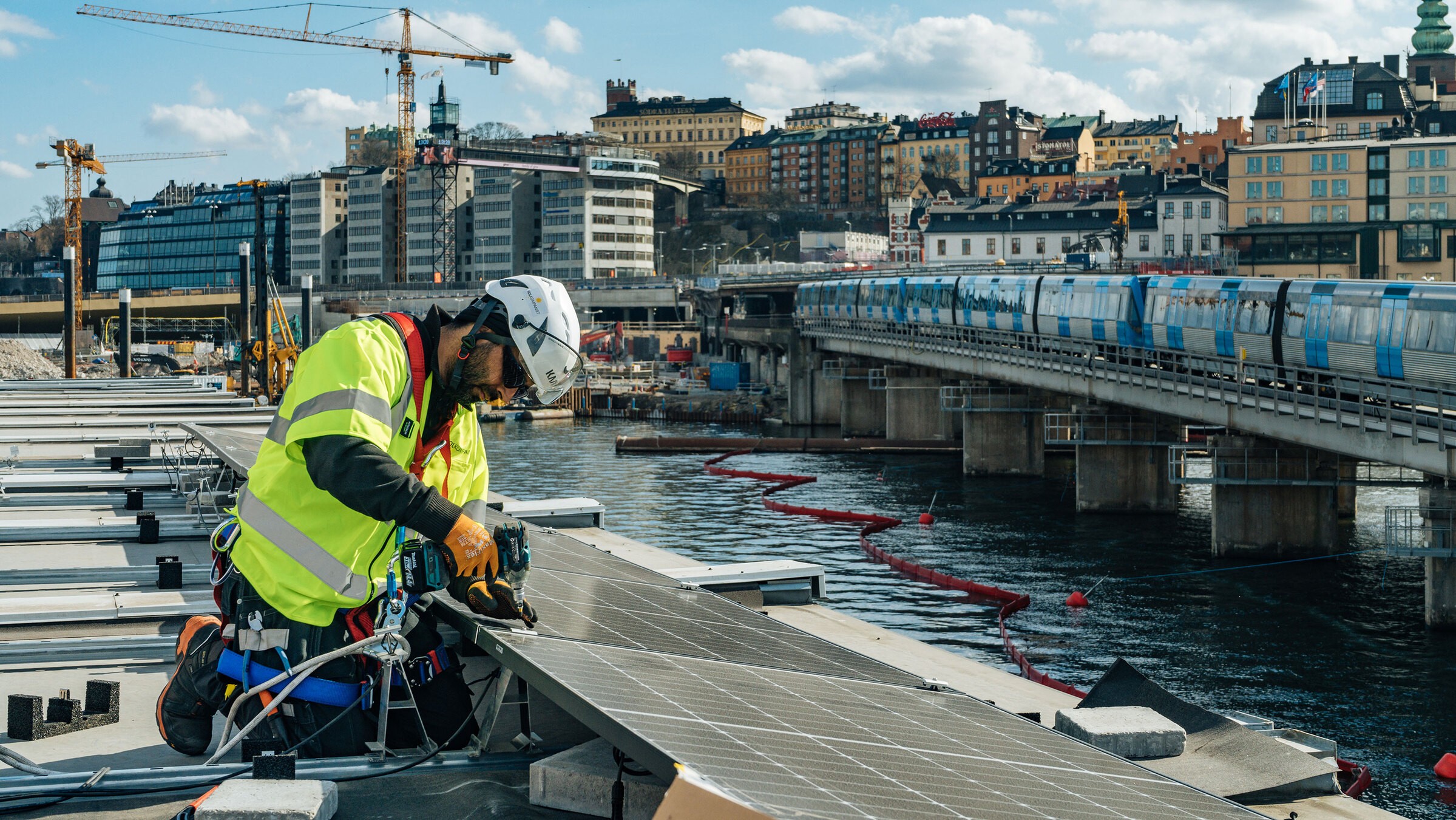 Skanska testar en ny lösning med solceller på byggbodarnas. Om försöket faller väl ut kan det bli en standardlösning för företagets alla etableringar inom ett par år. Arbetare installerar solpaneler på ett tak nära en flod i en urban byggmiljö.