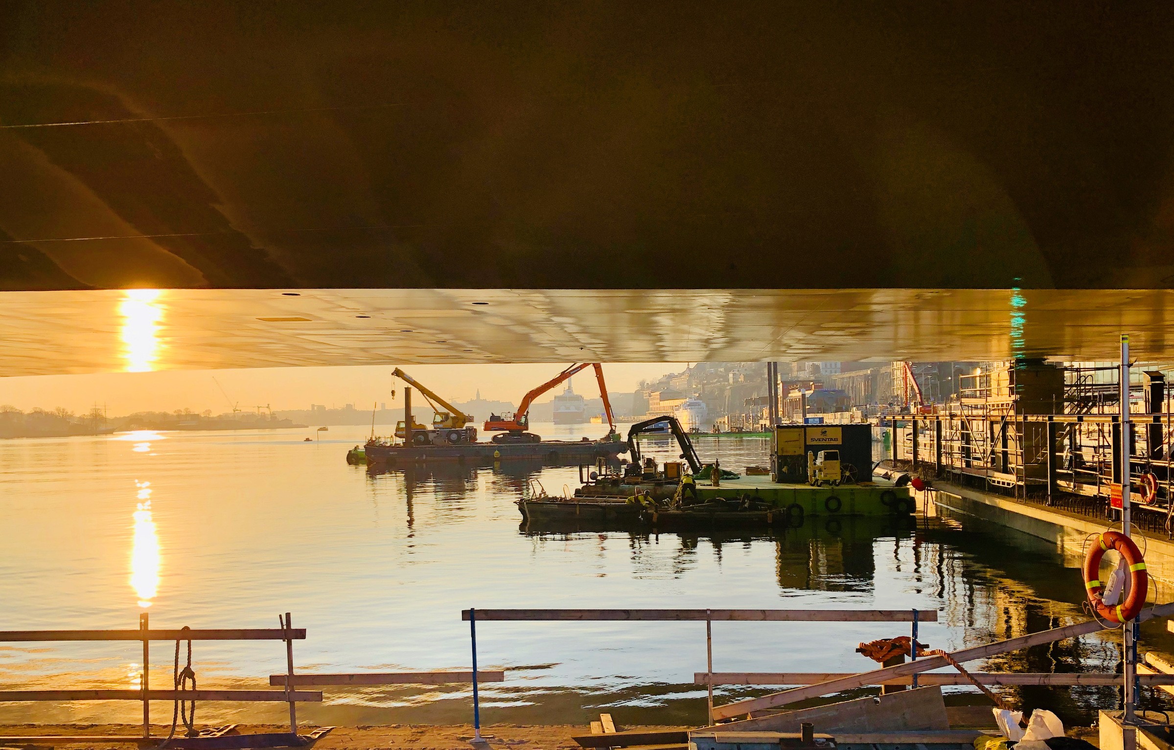 Slussenbron i morgonljus sedd underifrån. Byggutrustning vid en lugn strand vid solnedgången, reflekterad under en bro.