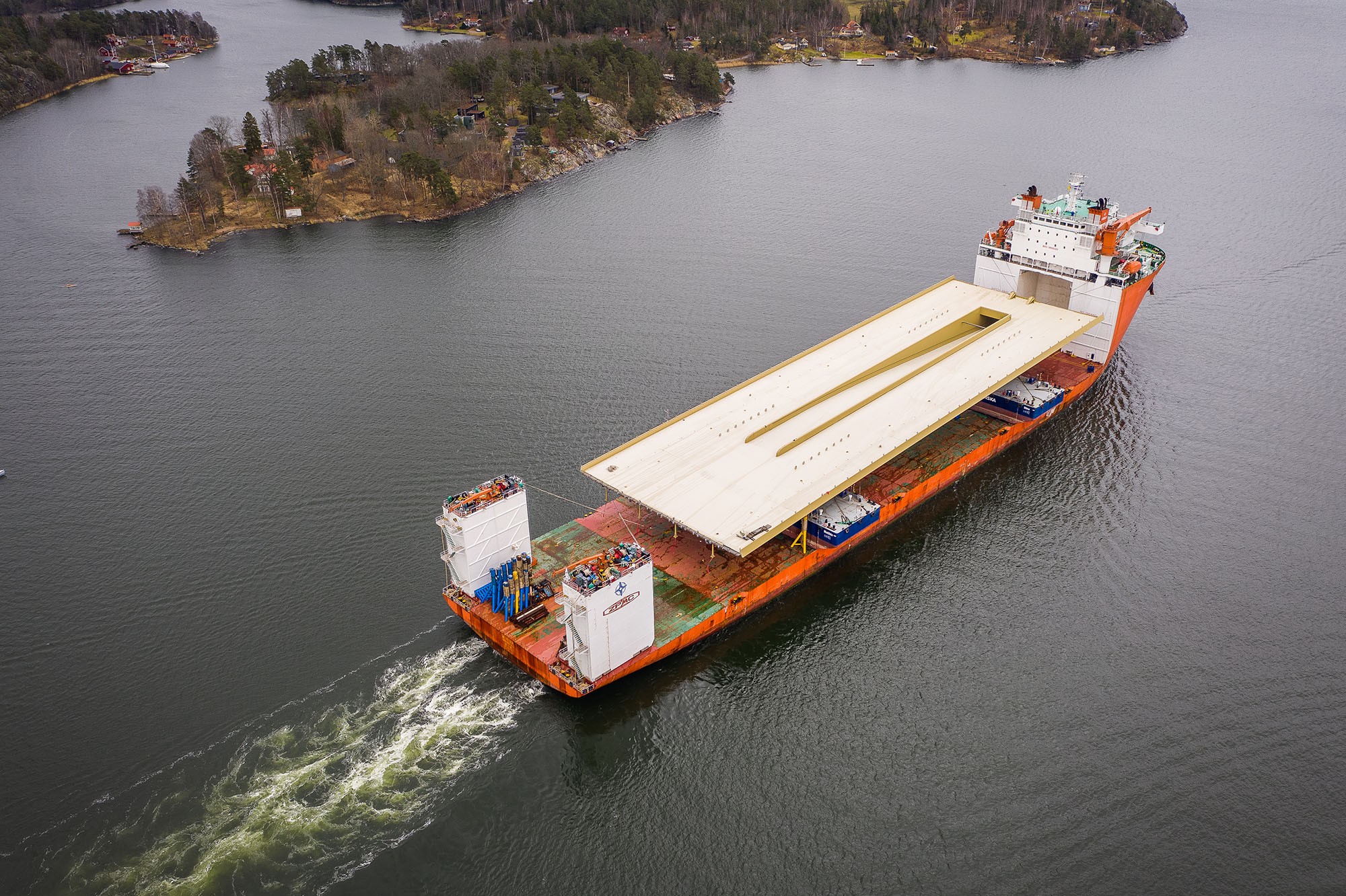 Slussenbrons färd genom skärgården i fågelperspektiv. Ett stort fraktfartyg transporterar en massiv struktur genom en naturskön vattenväg omgiven av öar.