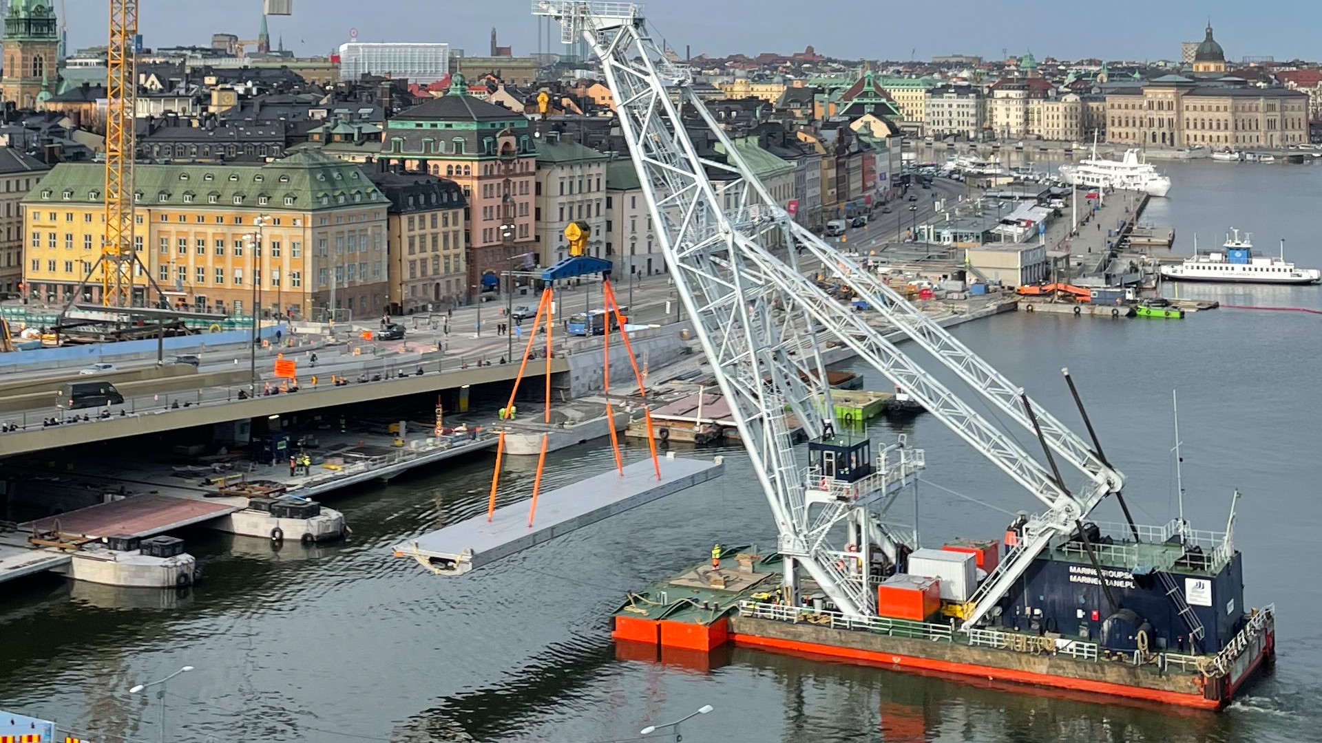 Slussens andra avbördningslucka transporteras på plats av Lodbrok. Den är 38 meter lång, 6,5 meter hög och väger 75 ton. Lyftkran på vattnet lyfter och transporterar en gigantisk lucka i stål på 75 ton. I bakgrunden syns byggprojektet Slussen i Stockholm.