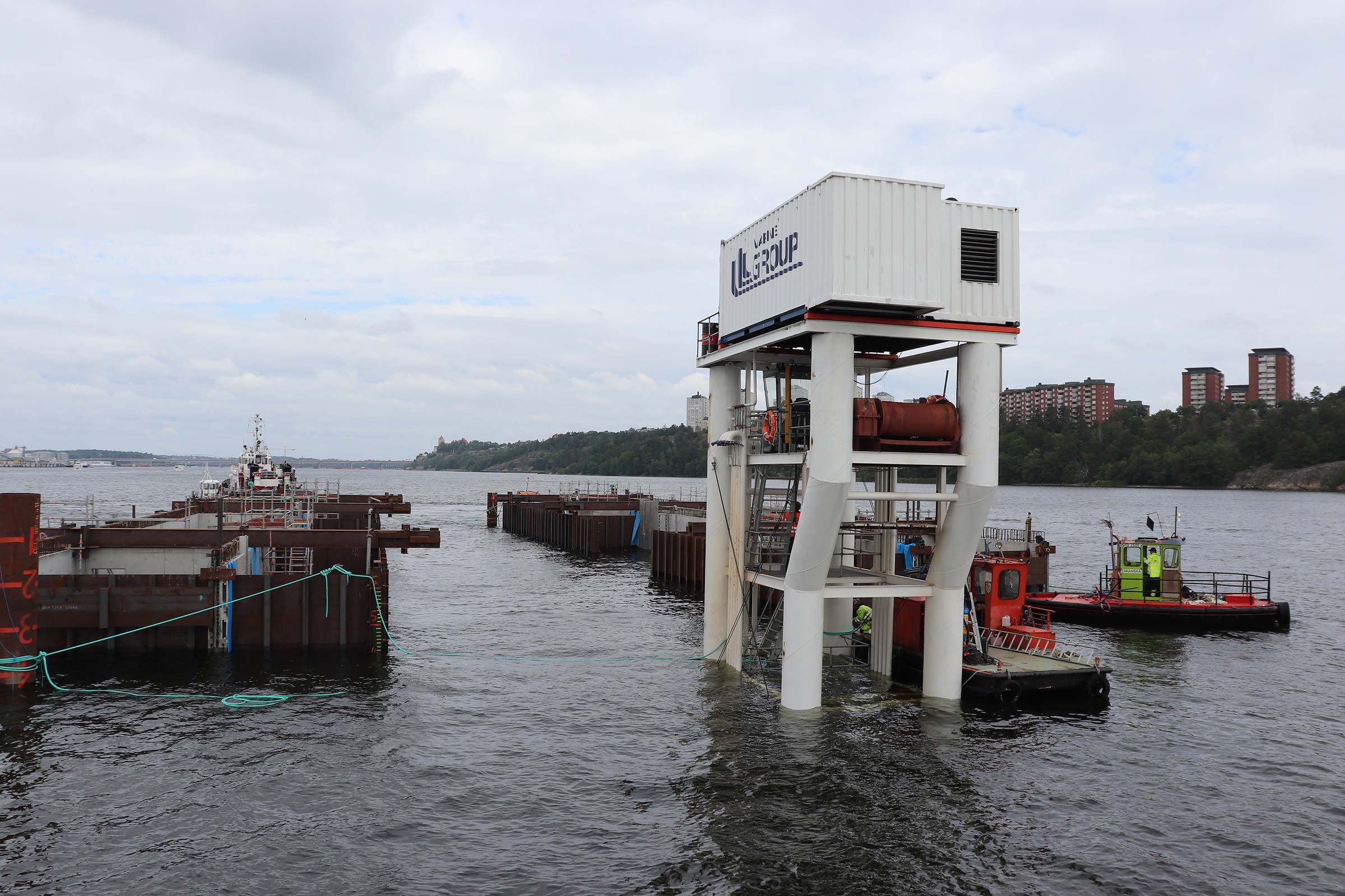 Slusskanalens delar transporteras på sänkpråm från Värtahamnen för sjösättning vid Loudden och vidare transport till Slussen-projektet. Kanalhalvorna sammanfogas på plats när de kommit till Slussens Mälarsida. Begränsningen är Centralbron samt tunnelbanebroarna därav transport av slusskanalen i två delar. Marin byggarbetsplats med en stor kran och pråmar på en flod, omgiven av stadens byggnader.