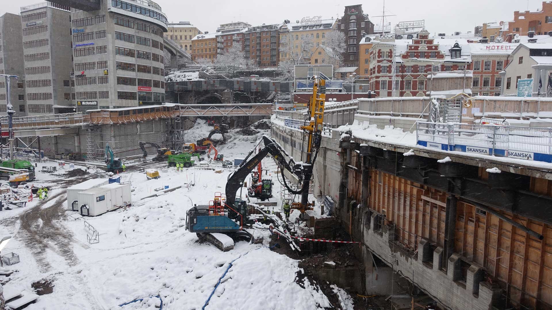 Södermalm under vintern Snötäckt byggarbetsplats med tunga maskiner i ett stadsområde omgivet av byggnader.