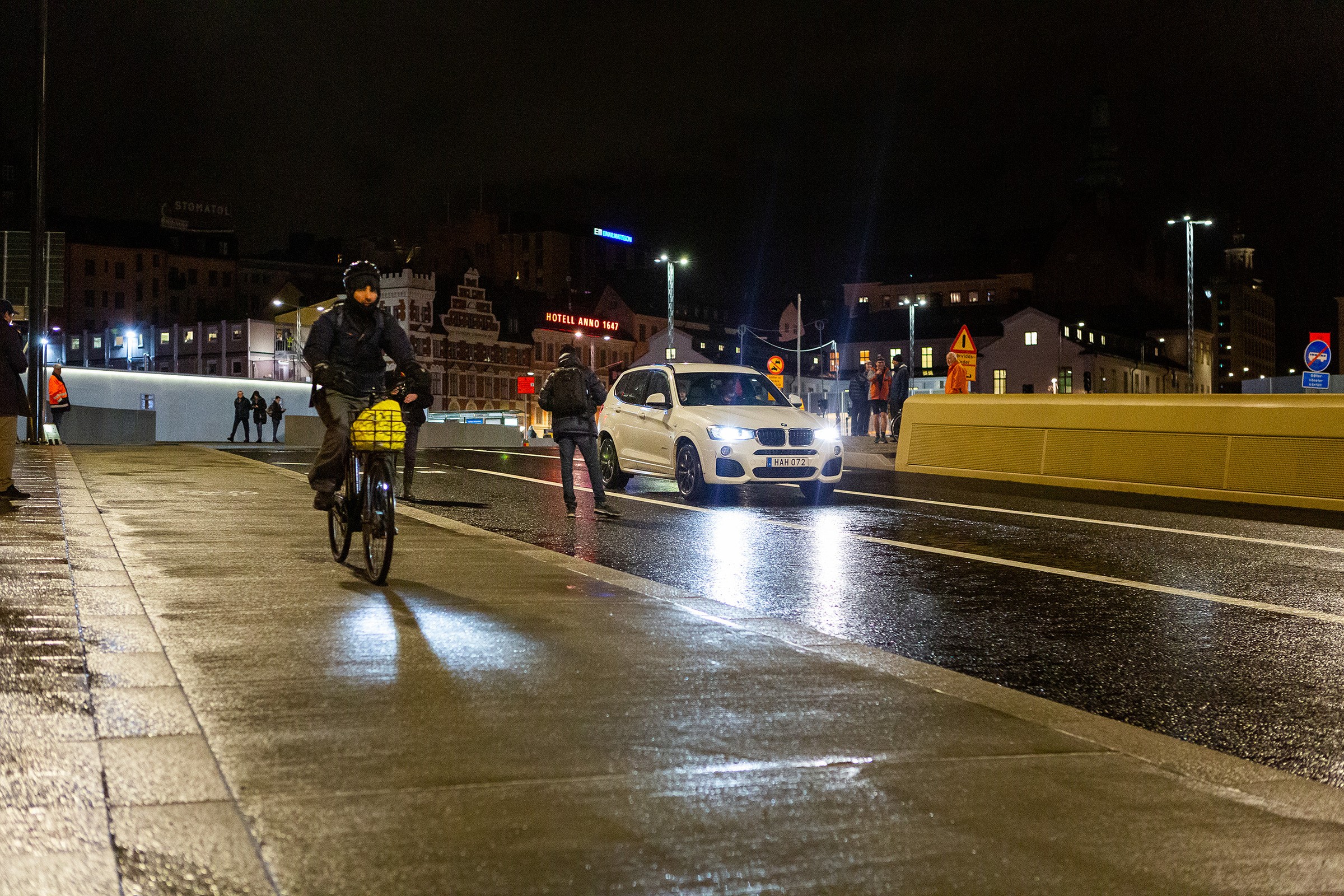 Trafiken släpptes på över nya Slussbron tidigt på morgonen den 25 oktober 2020. Cyklist och fotgängare navigerar en våt stadsgata på natten, med en vit bil som närmar sig.