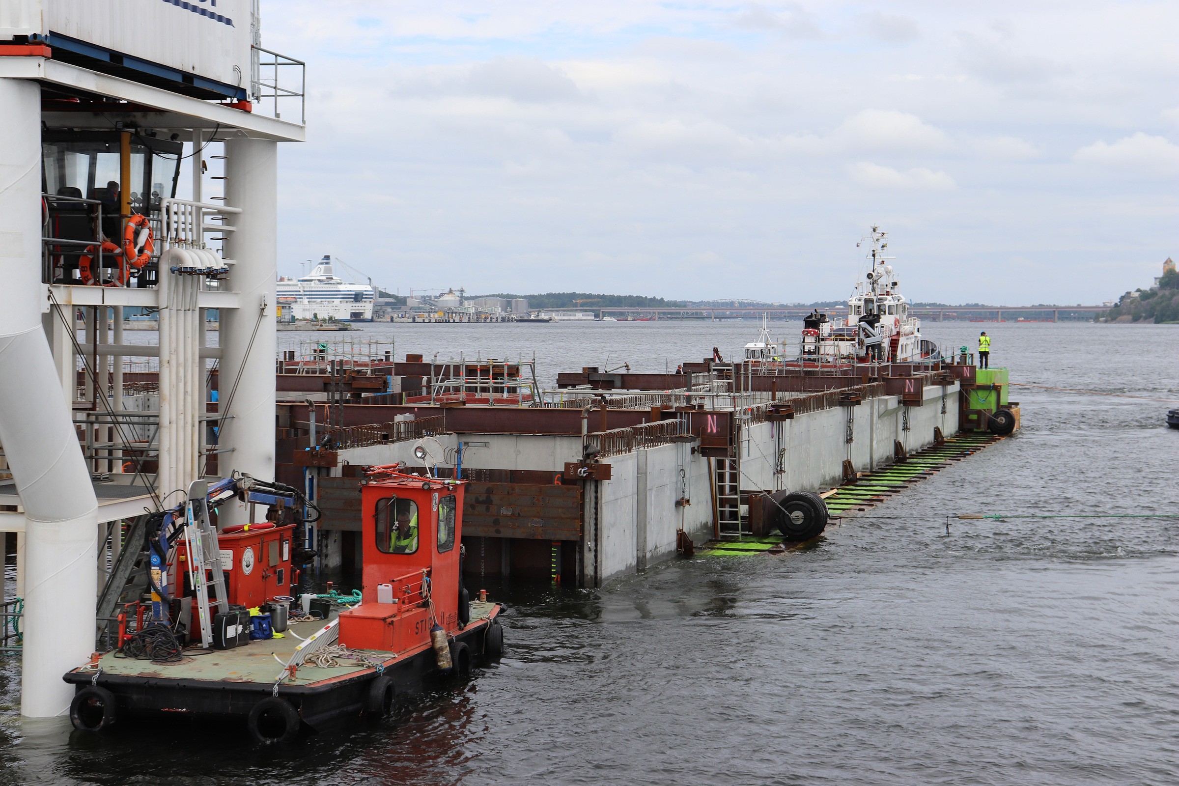 Transport av den västra delen av slusskanalen in till Stadsgårdskajen. Slusskanalen är delad i två längsgående delar för att möjliggöra senare intransport via Hammarbykanalen till Slussen på Mälarsidan. Enligt planen kommer båtar att kunna slussa genom den nya slusskanalen från 2024. Byggnation av kaj med pråmar och bogserbåtar på en flod, med industriell utrustning och fartyg.