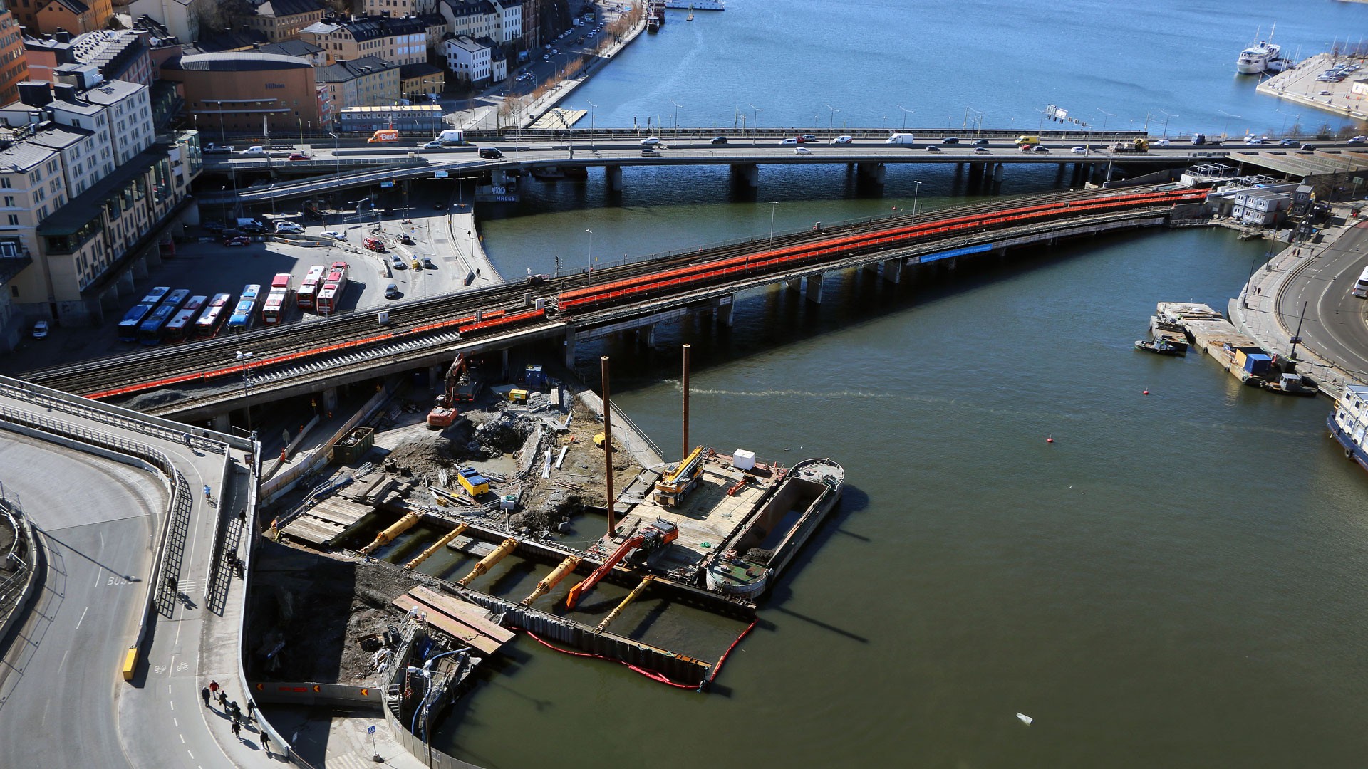 Vy inåt Mälaren från den mäktiga tornkranen på Slussen. Flygvy över brokonstruktion över en flod, med synliga kranar och omgivande stadsmiljö.
