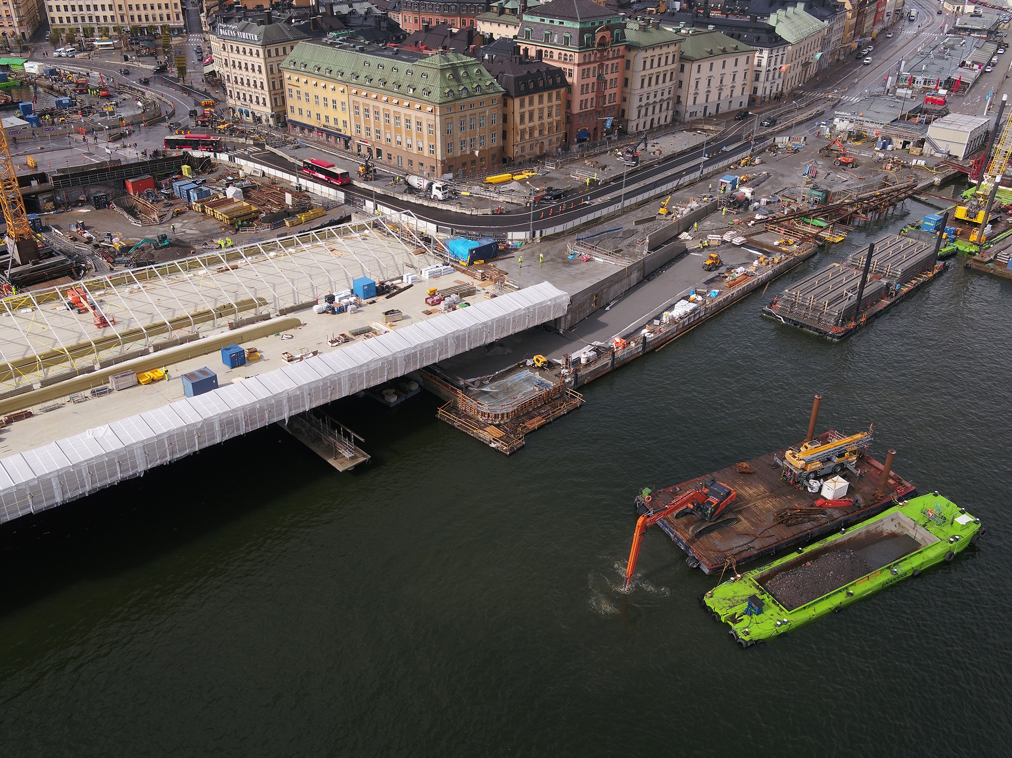 Vy över brofästet mot Gamla stan. Flygvy över byggarbetsplats vid stadens vattenfront med broarbete och maskiner i arbete.