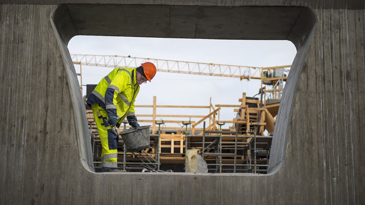 Byggnadsarbetare i högsynlighetsutrustning håller en hink, inramad av en betongöppning.