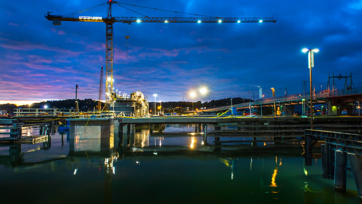 En byggkran lyser upp en brygga i skymningen, reflekterande på lugnt vatten under livfulla himlar.