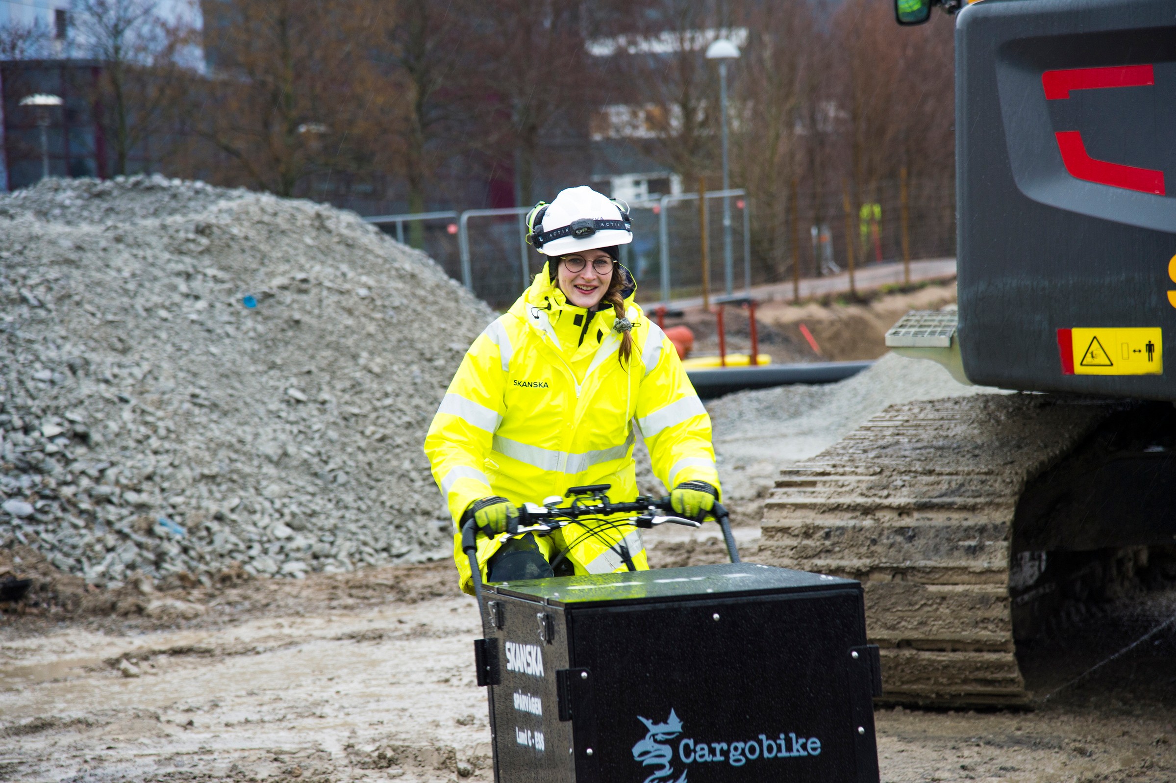 Byggnadsarbetare i högsynlighetsutrustning kör en Cargobike nära en grushög på platsen.