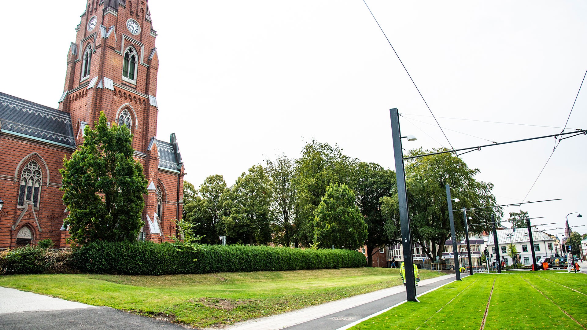 Skanska har byggt en ny spårväg mellan Lunds Central och ESS. Historisk röd tegelkyrka med hög spira bredvid en modern spårvagnslinje i en grön stadsnära miljö.