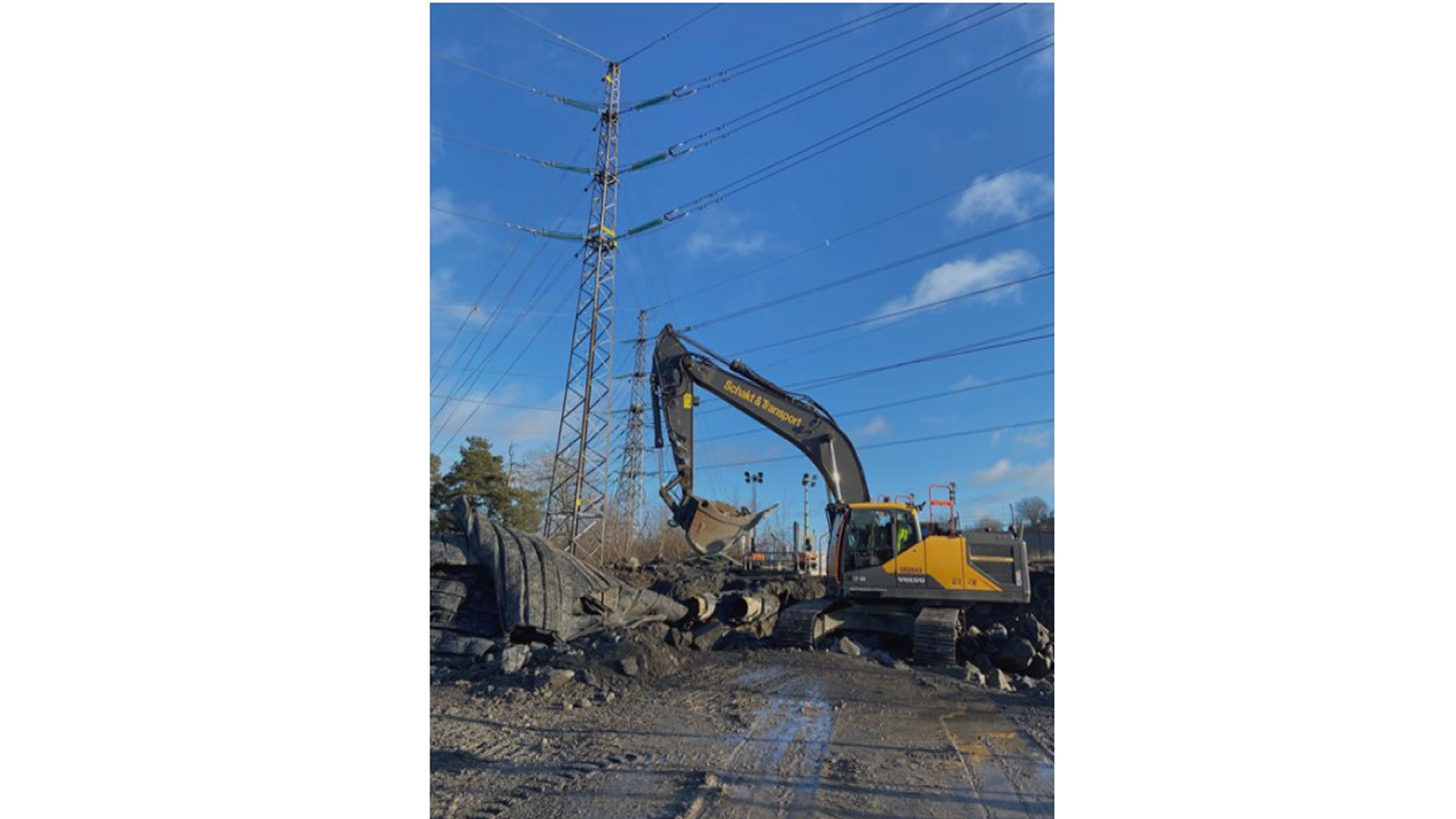 Arbete under befintliga luftledningar har ställt höga krav på säkerheten. Grävmaskin bryter asfalt nära ett kraftledningstorn under en klarblå himmel på en byggarbetsplats.