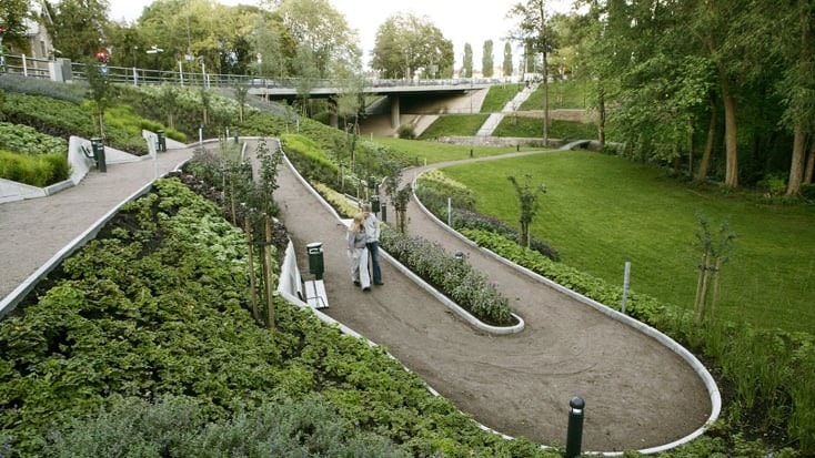 En fridfull parkstig med frodig grönska, med ett par som promenerar under en bro.