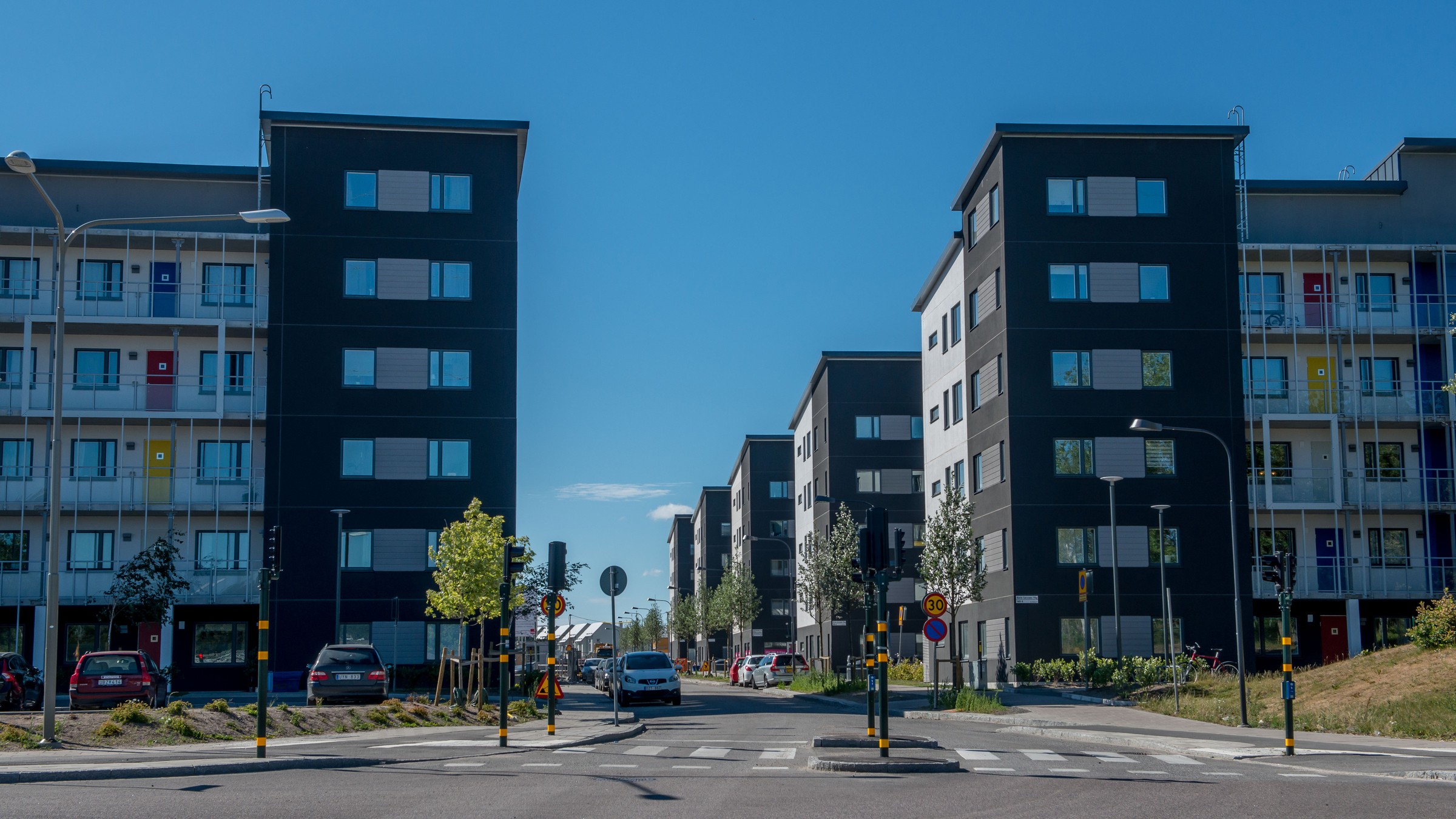 Moderna lägenhetsbyggnader flankerar en gata under klarblå himmel, med parkerade bilar och träd.