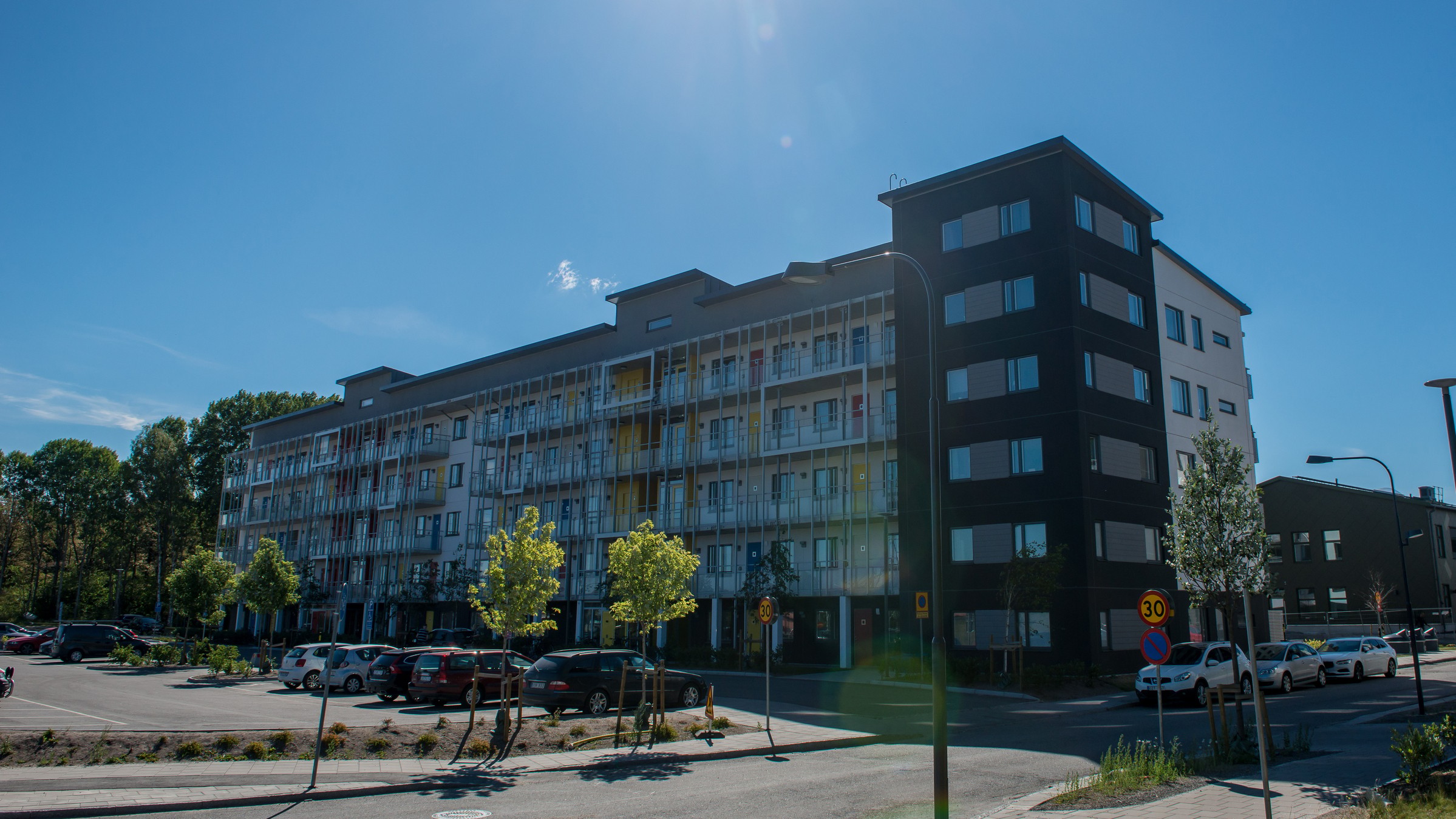 Modern lägenhetsbyggnad under en klar blå himmel med parkerade bilar och träd i förgrunden.