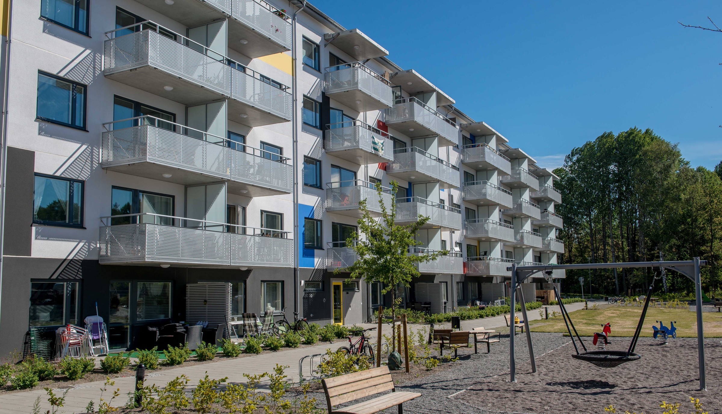 Modern lägenhetsbyggnad med balkonger, lekplats med gungor, bänkar och anlagd trädgård.