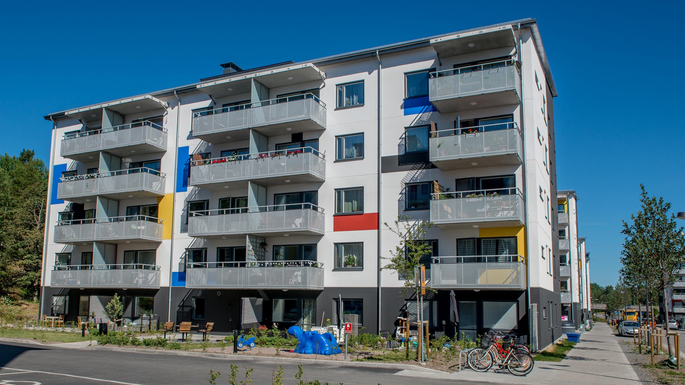 Modern lägenhetsbyggnad med färgglada inslag, balkonger och omgivande grönska under en klar himmel.