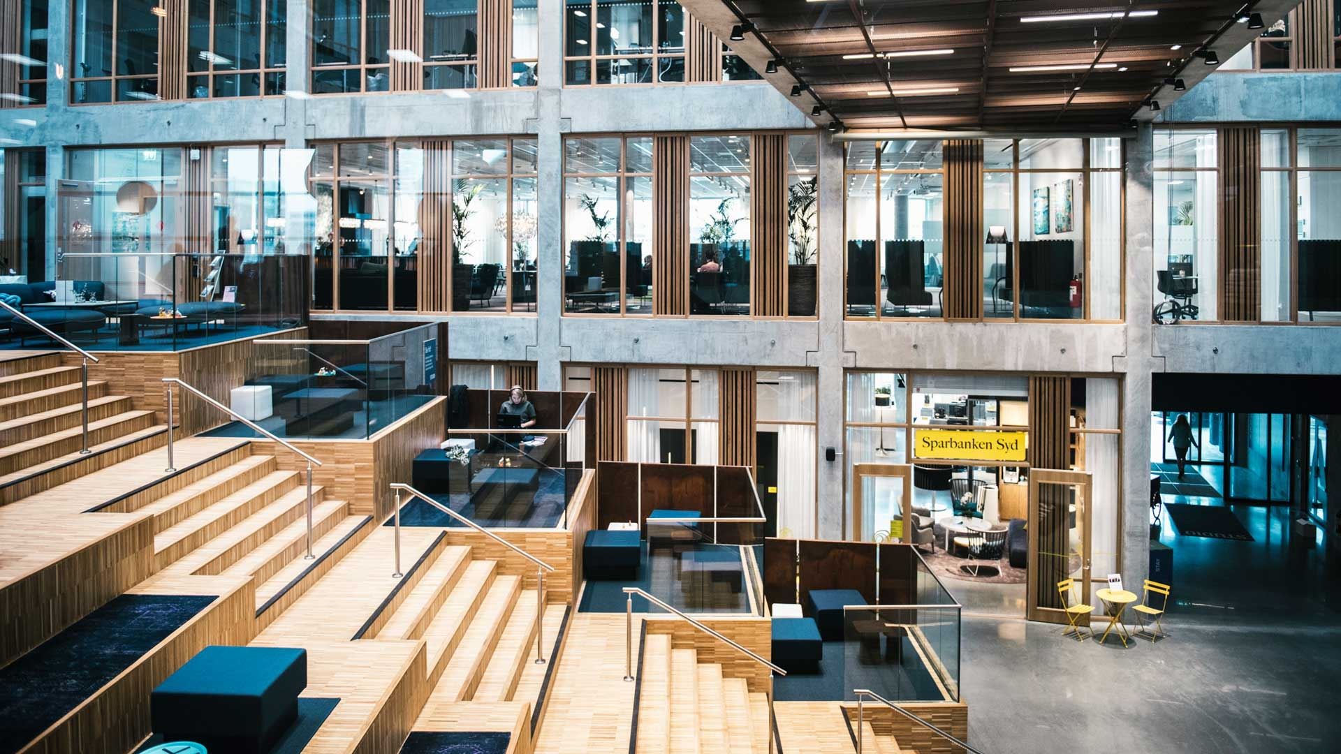 Atrium Modern kontorsinteriör med öppet utrymme, trappstegsformade sittplatser, glasavdelare och synliga arbetsstationer.
