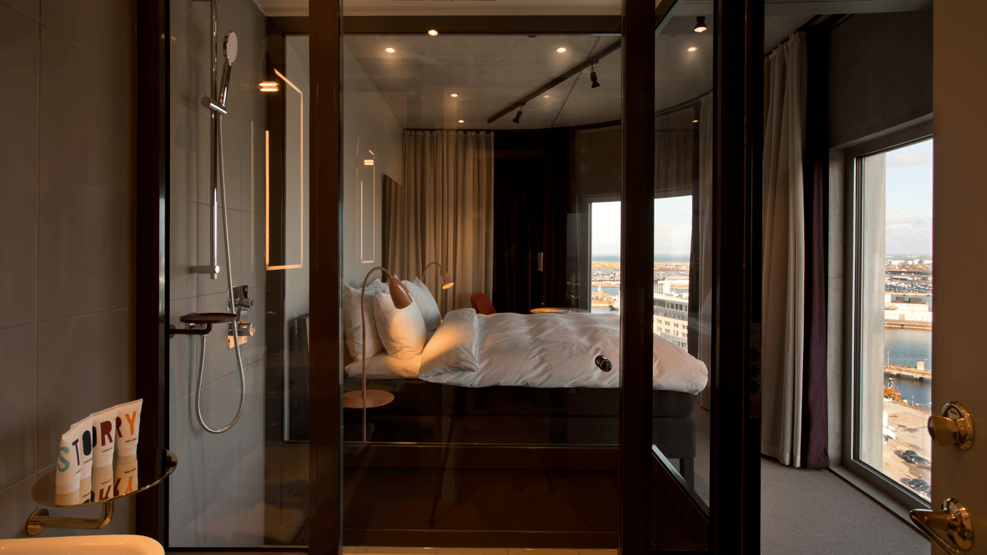 Story Hotel Modern hotellrum med glasväggad dusch, mysig säng och stadsvy genom stora fönster.