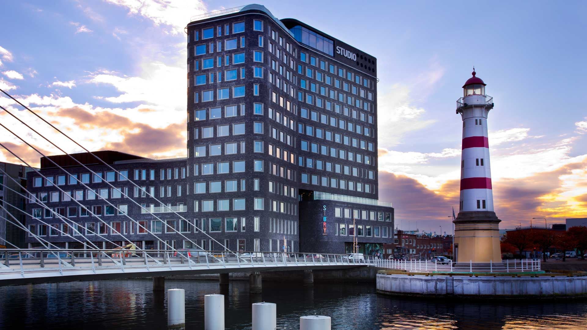 Studio kvällsbild Modernt hotell vid vattnet med fyr och bro under en färgglad solnedgångshimmel.