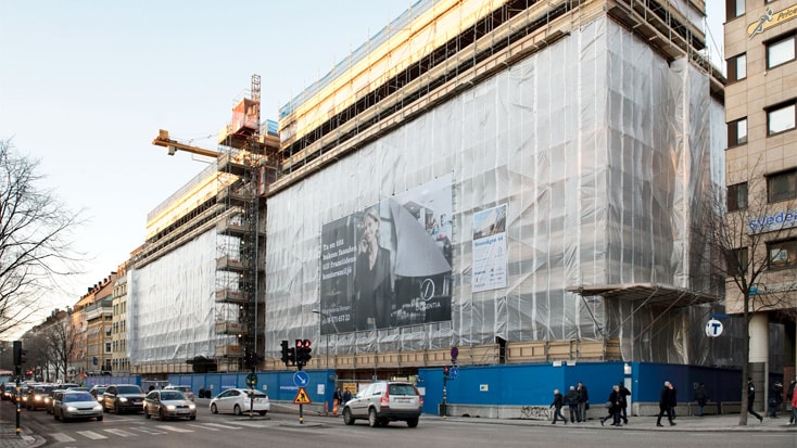 Byggnad under konstruktion med byggnadsställningar och skyddstäckning på en livlig gatukorsning i staden.