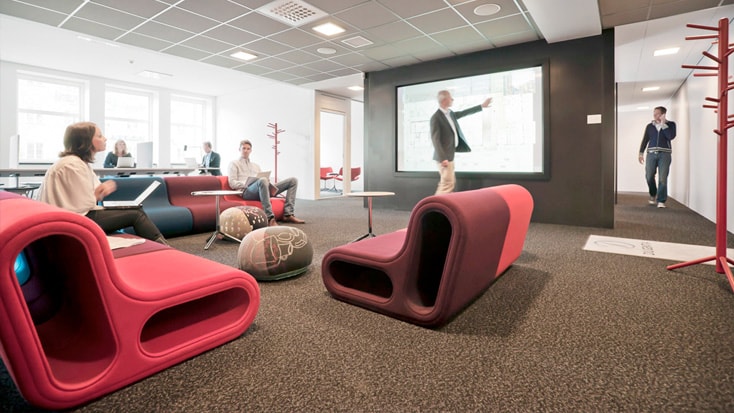Modern kontorslounge med färgglada sittplatser, människor som arbetar och en presentationsskärm i bruk.