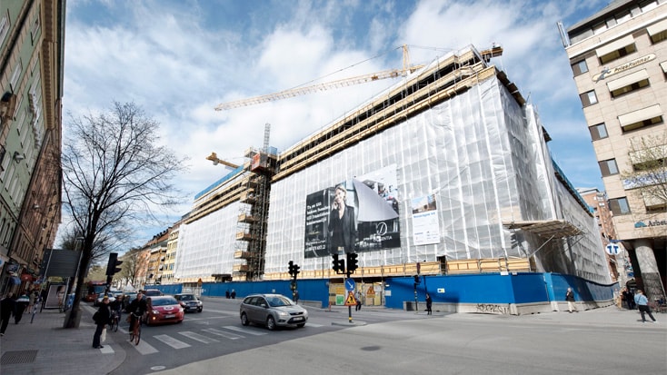 Stadsbyggnadsplats med kranar, täckt byggnad och gatutrafik vid en livlig korsning.