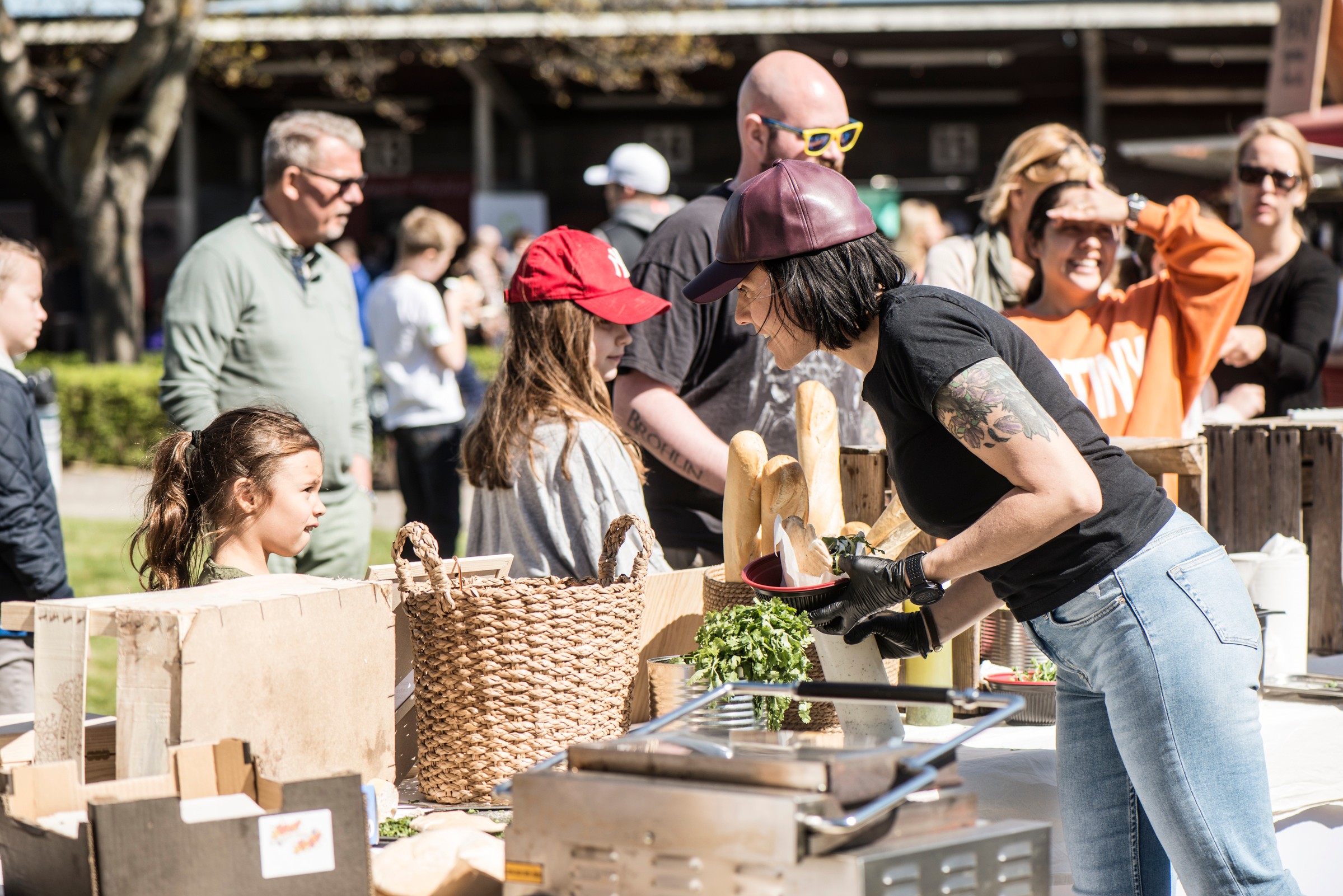 Food markets och andra aktiviter året runt gör Täby Park till en mänsklig stadsdel med rikt folkliv. Utomhusmatmarknad med kvinna som serverar bröd och grönsaker till en folkmassa, solig dagsstämning.