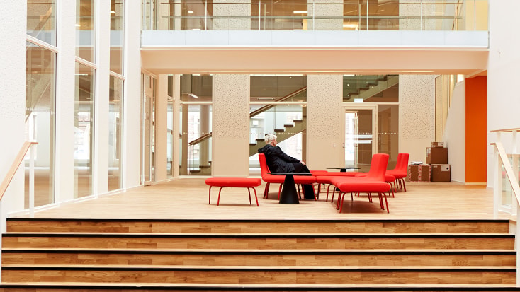 Gångstråk och broförbindelser skapar dynamik i huset Modernt atrium med röda sittplatser, trägolv och en person som sitter mitt i stark belysning.