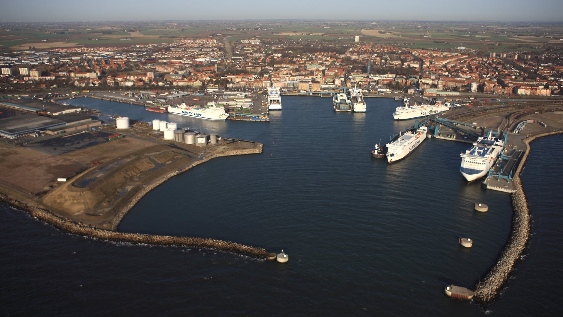 Flygvy över en livlig kusthamn med flera fartyg förtöjda och en stad i bakgrunden.