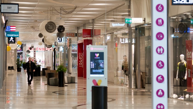 Modern shoppingmallskorridor med butiker, digital kiosk och vägbeskrivningar för besökare.