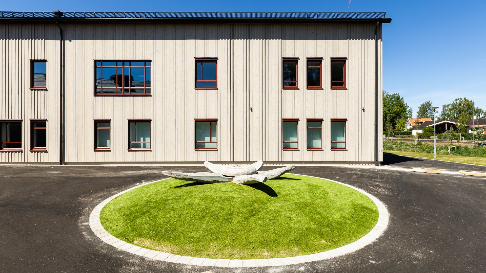 Exteriör av Västerängsskolan i Nybro med träfasad i ljus furu och röda fönster, samt konstverk på skolgården.