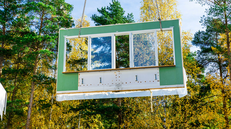 En prefabricerad väggsektion med ett stort fönster lyfts av en kran i ett skogsområde.